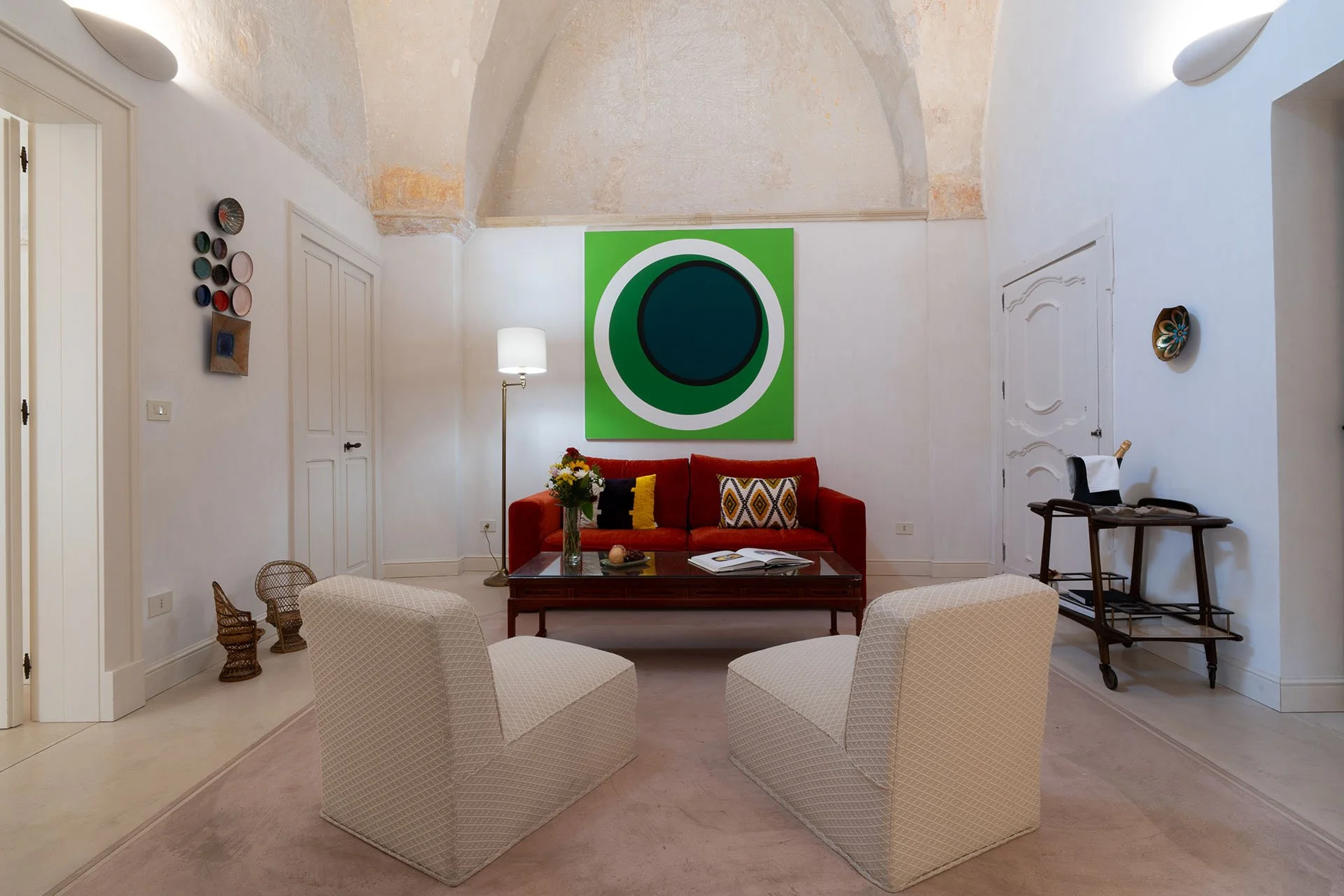A living room with white walls, a rust-colored sofa with patterned pillows, two white armchairs, a dark wooden coffee table, a vase of flowers, a floor lamp, and colorful wall art. There is a large green and blue abstract painting above the sofa.