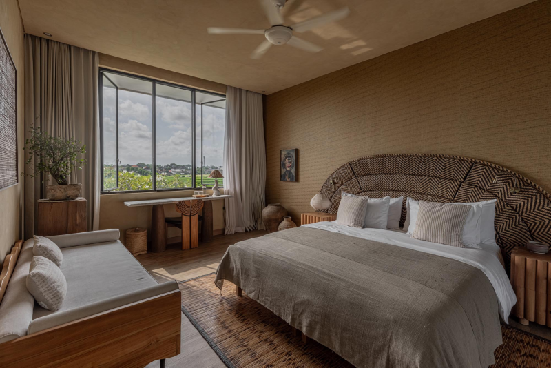Well-decorated bedroom with a large bed, pillows, and a curved headboard, a daybed with cushions, a large window with curtains, and minimalist wooden furniture in neutral tones.