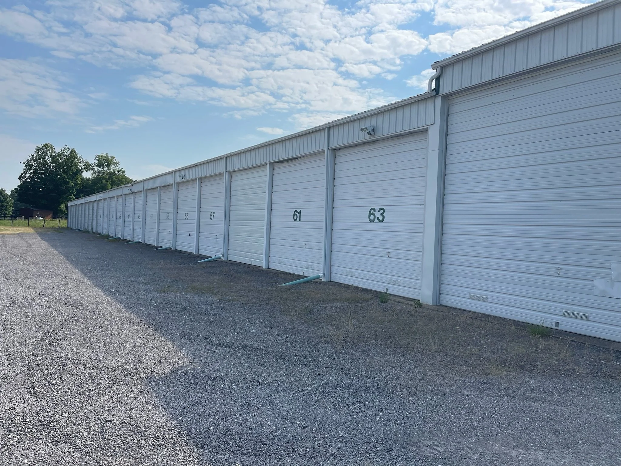 Row of white storage units with numbered doors, and a gravel parking lot.