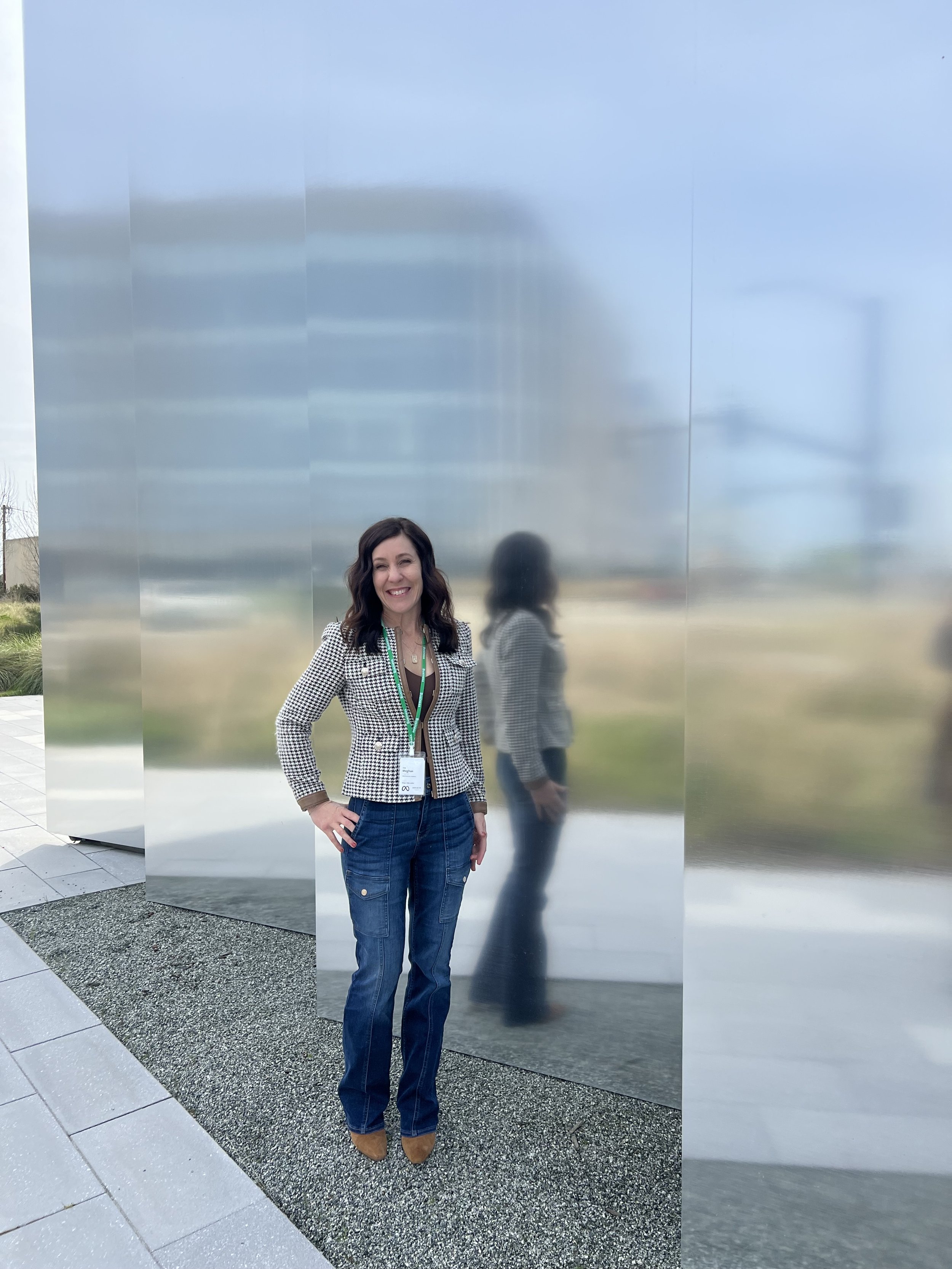 A woman with dark hair wearing a black and white houndstooth blazer, black top, blue jeans, and brown shoes standing outside next to a reflective metallic wall, smiling at the camera.