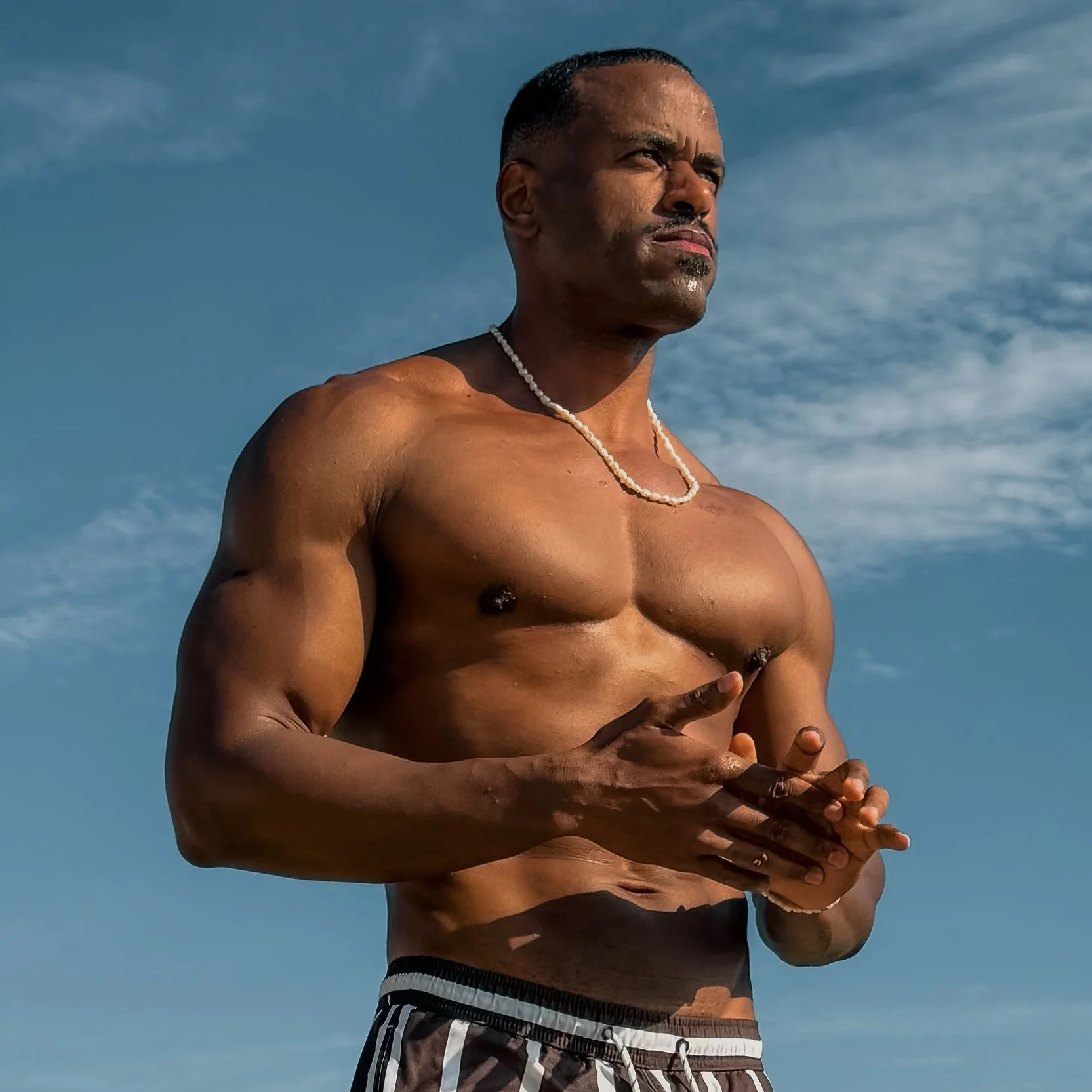 	
Renan Soulza criador de conteúdo musculoso e sem camisa, com colar de conchas, em pose relaxada sob céu azul com nuvens, mãos juntas na frente.