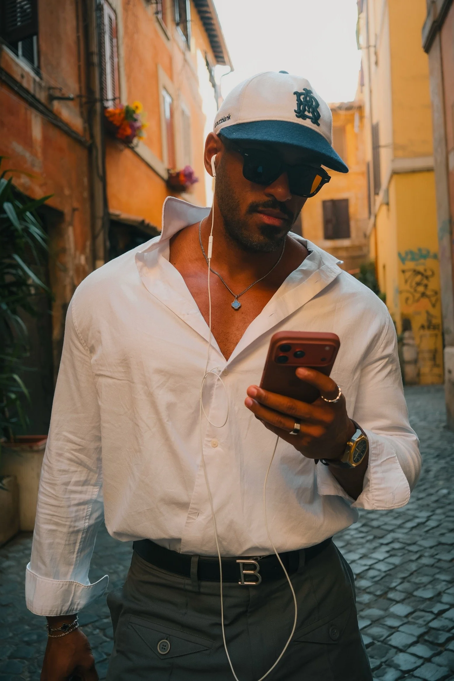 	
Renan Soulza criador de conteúdo de óculos escuros, boné branco e camiseta branca, usando fones de ouvido, olhando para o celular, andando por uma rua de paralelepípedos em uma área urbana com edifícios coloridos ao fundo.
