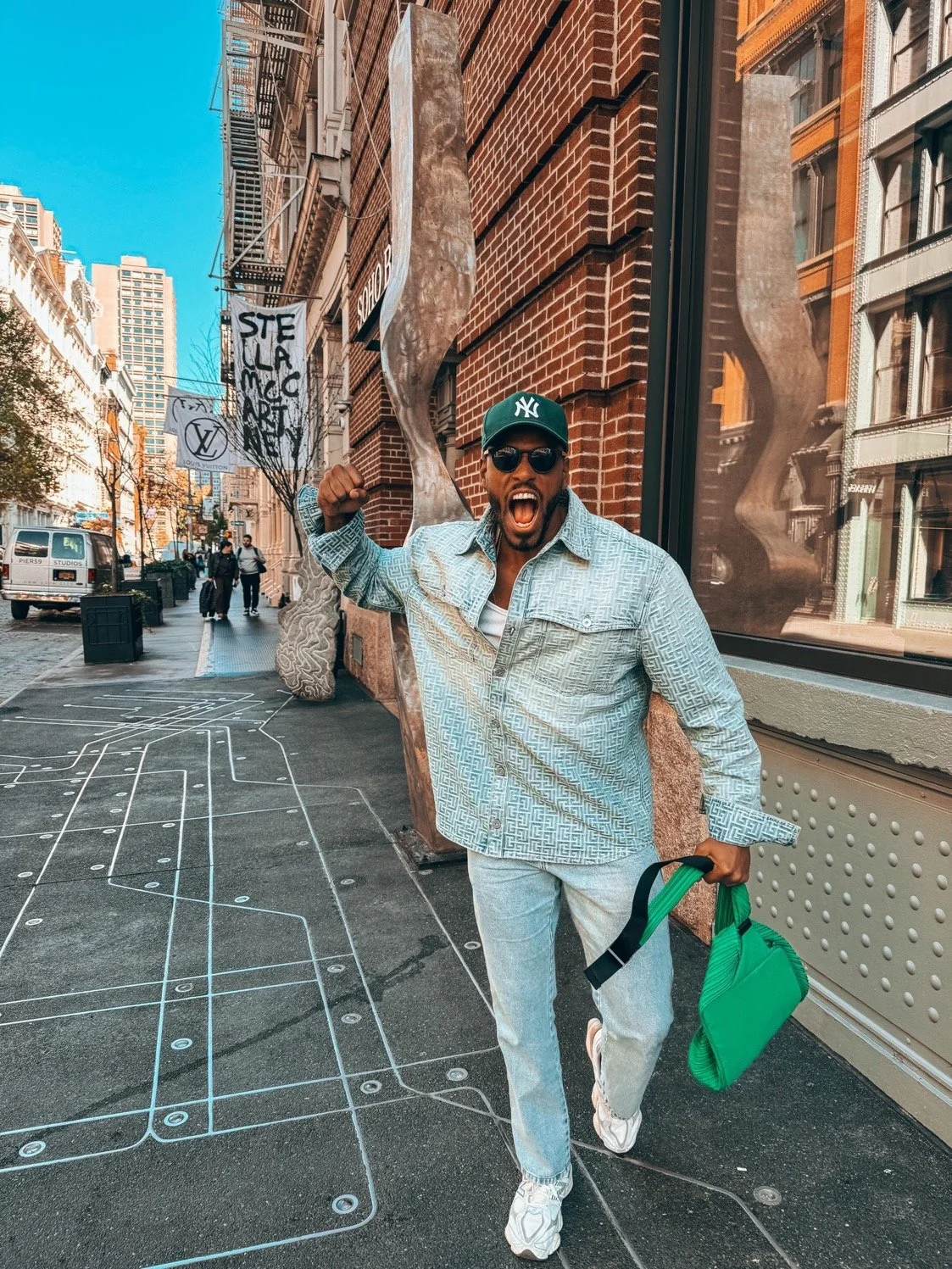 Renan Soulza agitando um braço com expressão de entusiasmo na rua de SoHO em NY, usando boné do Yankees, óculos escuros, camisa da Pierre B, mochila verde, calça jeans clara e tênis brancos, ao lado de uma escultura moderna de metal enorme e paredes.