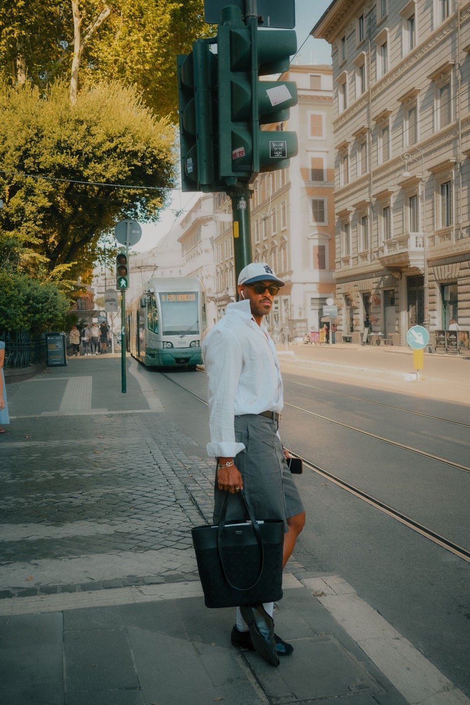	
Renan Soulza criador de conteúdo com roupas sociais, óculos escuros e chapéu, carregando uma bolsa grande preta e um celular, aguardando para atravessar a rua em uma cidade urbana.