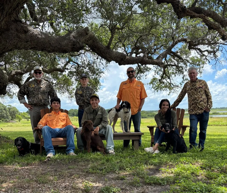 A diverse group of seven people, including young adults and older adults, along with five dogs, pose outdoors under a large tree with a scenic landscape of grass, trees, and water in the background. Some individuals are seated on chairs, while others stand, all casually dressed.