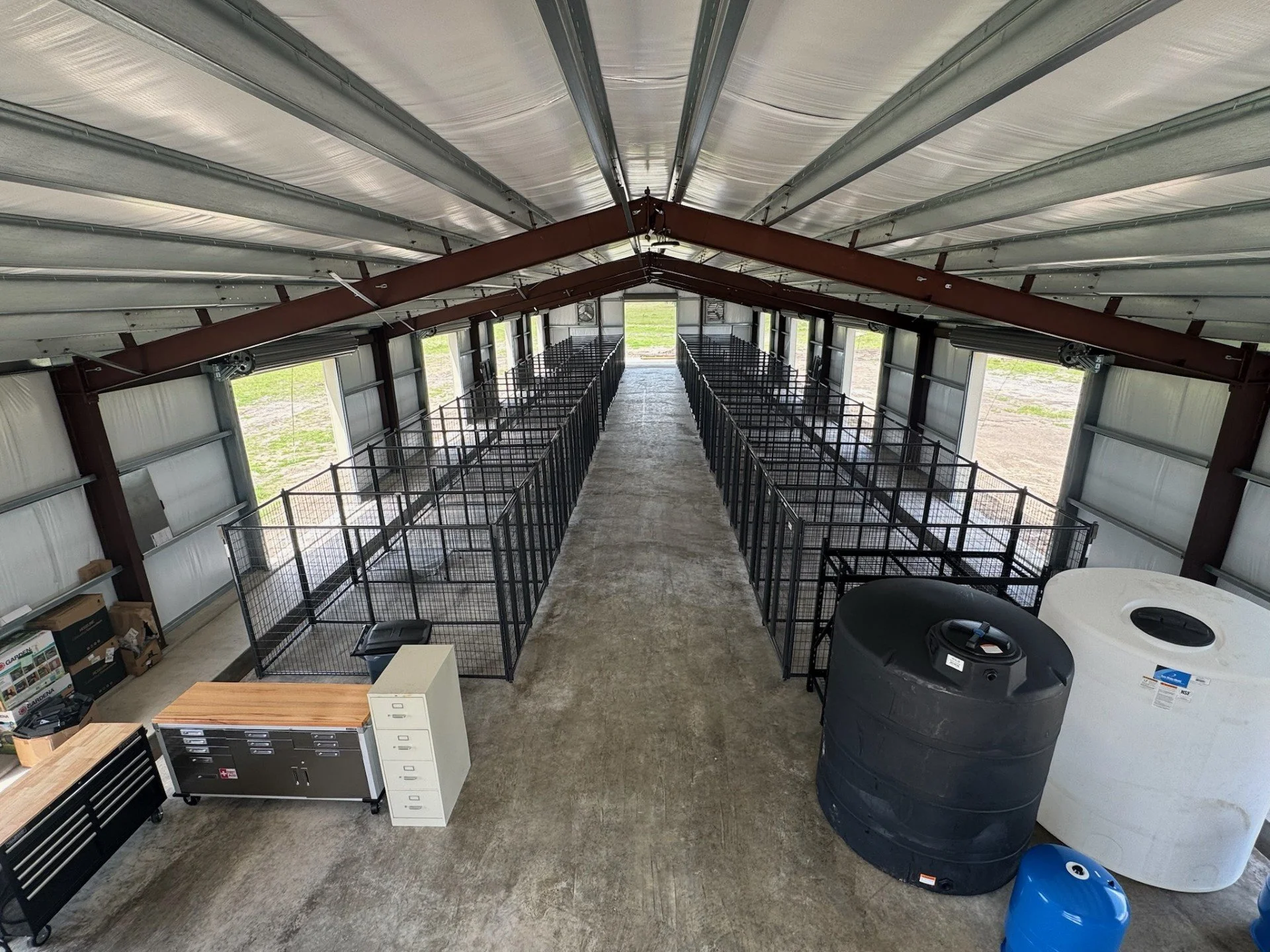 Large indoor area with metal animal enclosures arranged in two rows, water tanks, tool chests, and storage containers, likely used for livestock or pet housing.