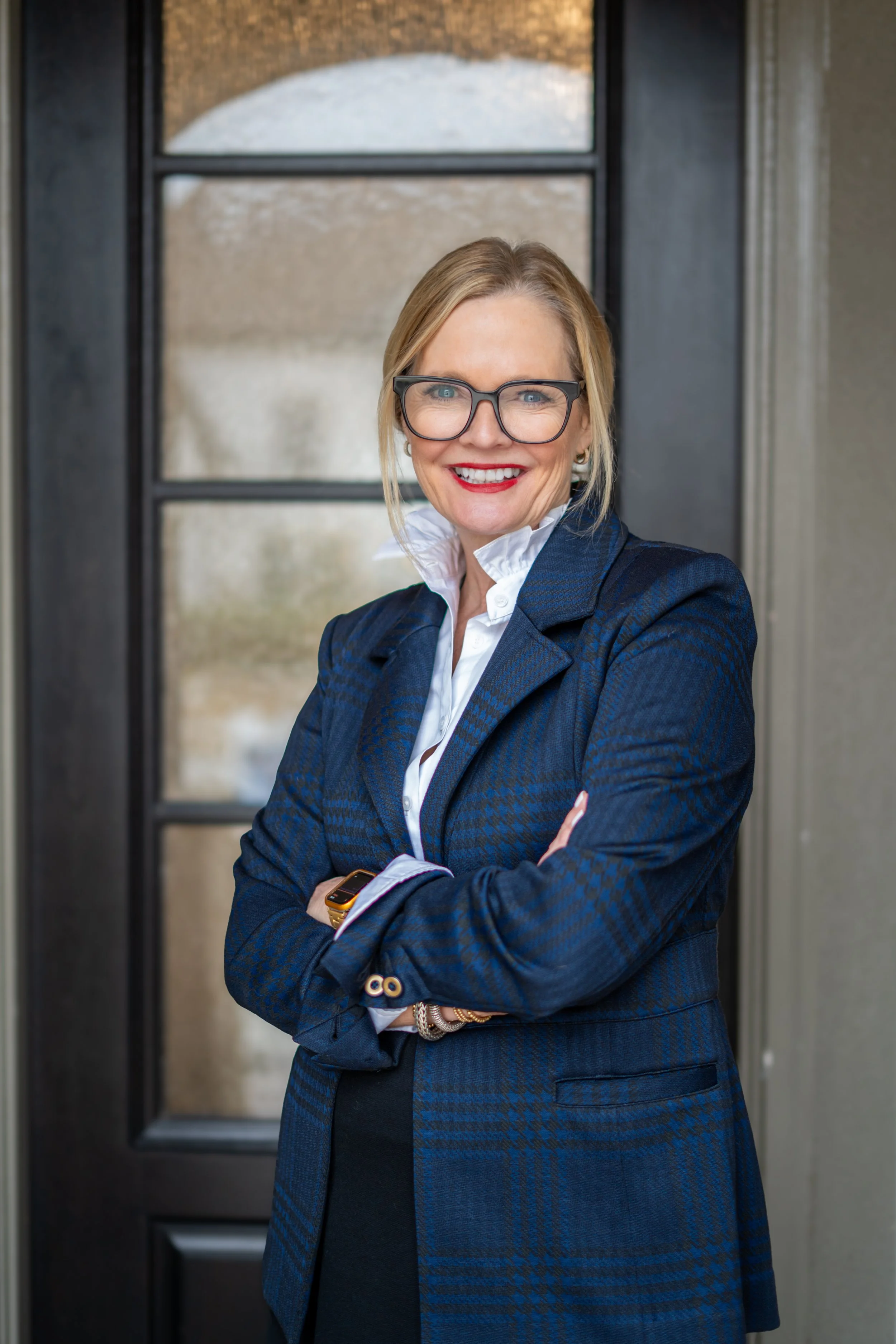 A smiling woman with glasses standing in front of a door, dressed in a blue blazer and white shirt.