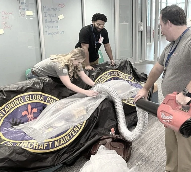 People working on the maintenance of a large aircraft engine, covered with a black tarp with a logo, in a bright indoor space.
