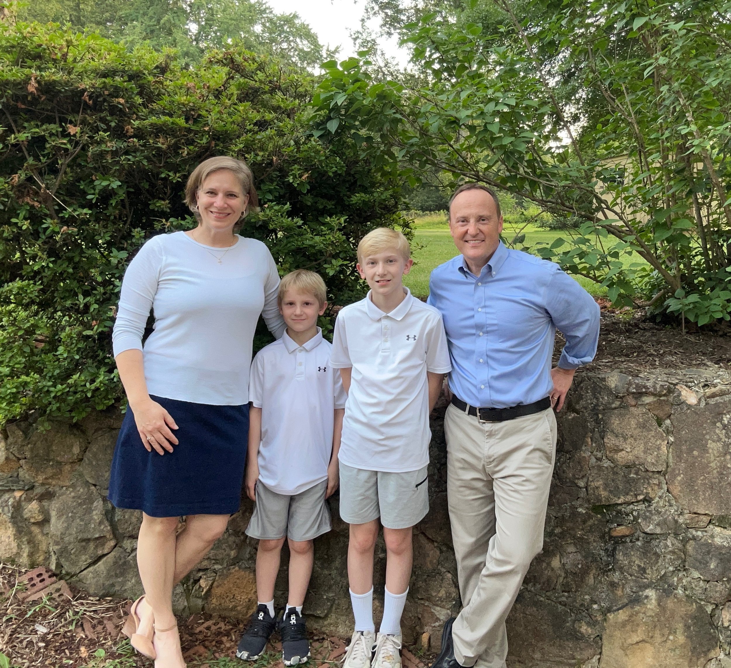 Family of five standing outdoors in front of lush greenery and a stone wall. Two adult women and three boys are smiling at the camera. The woman on the left is wearing a white top and navy skirt; the woman on the right is in a light blue shirt and kh