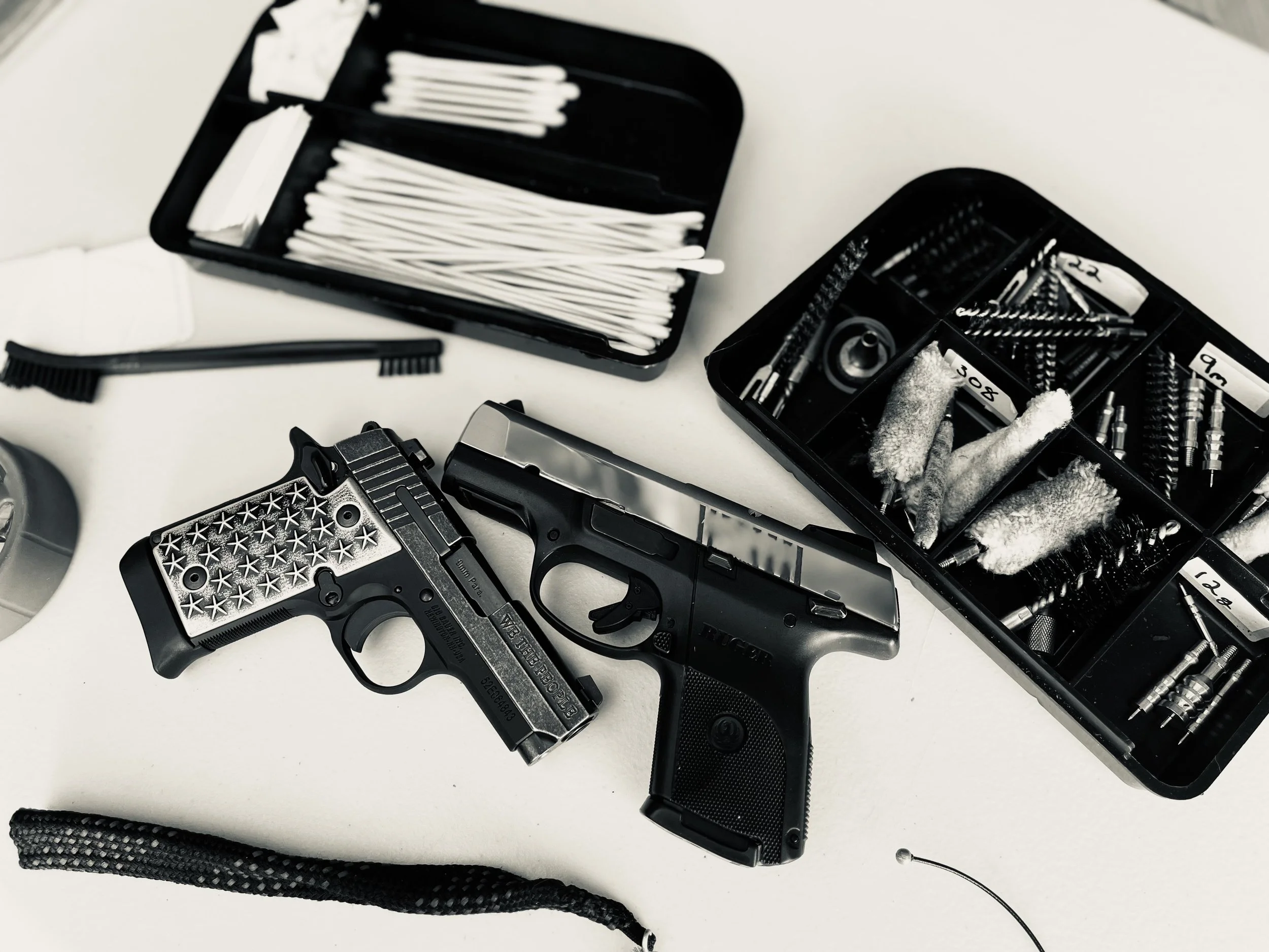 Two handguns, one with a star-patterned grip, placed on a white surface surrounded by gun cleaning supplies in black organizers, with cleaning cloths, brushes, and small parts.