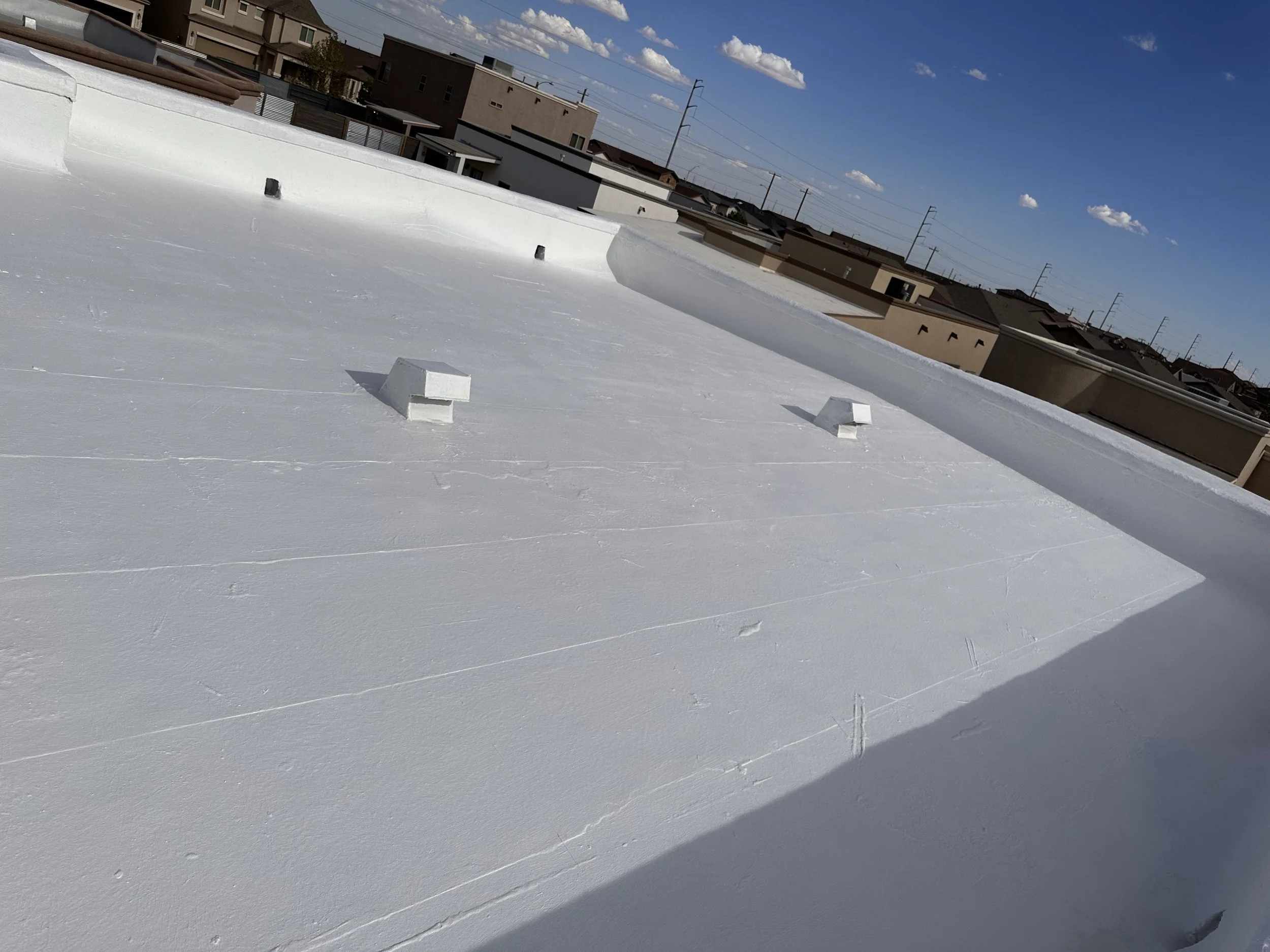 View of a flat white roof with three small vents or exhaust pipes, under a blue sky with some clouds, and surrounded by neighboring buildings and utility poles and wires.