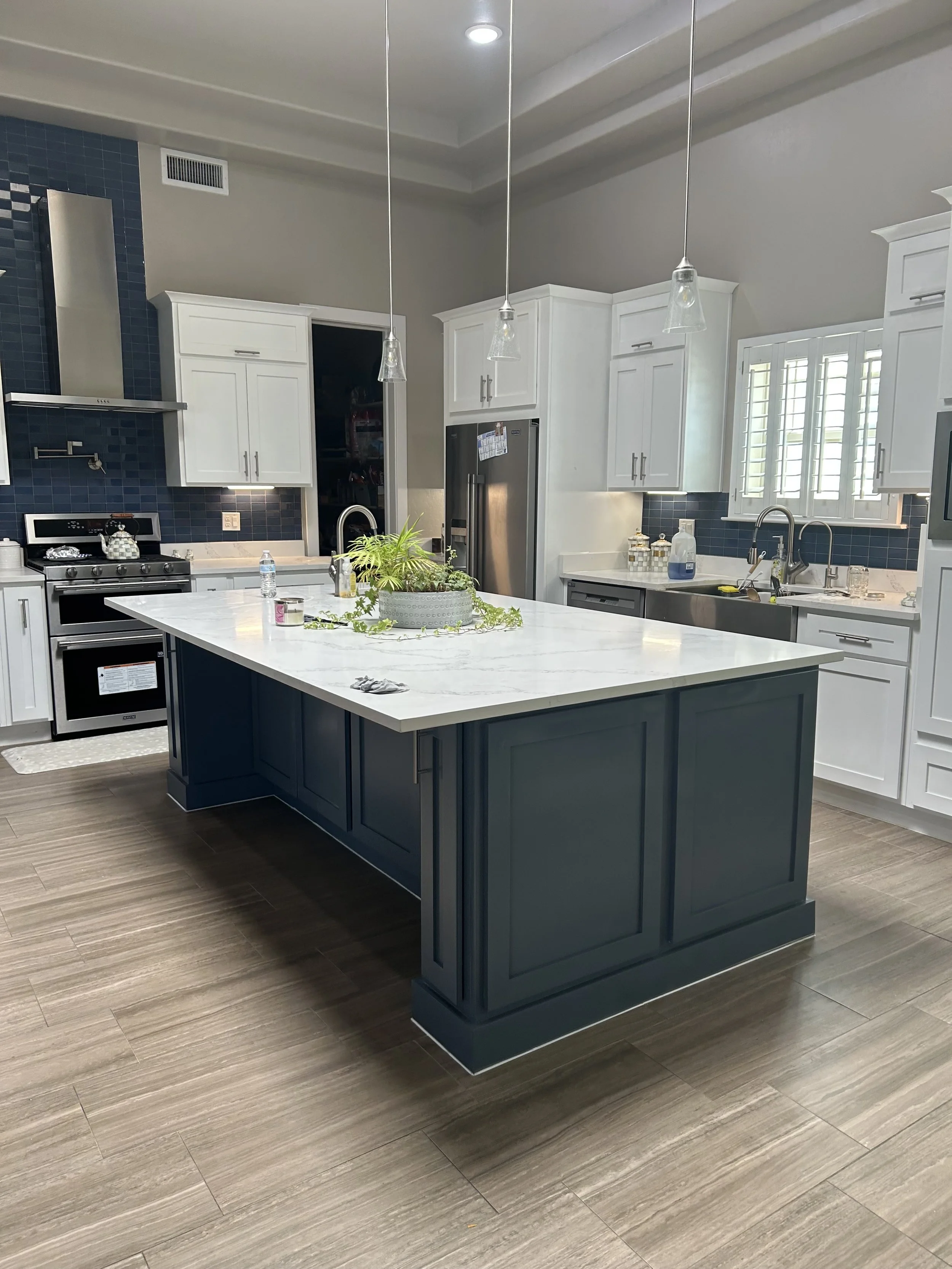 Modern kitchen with white cabinets, a navy blue island, stainless steel appliances, dark blue backsplash, wood-look flooring, and a window with white shutters.