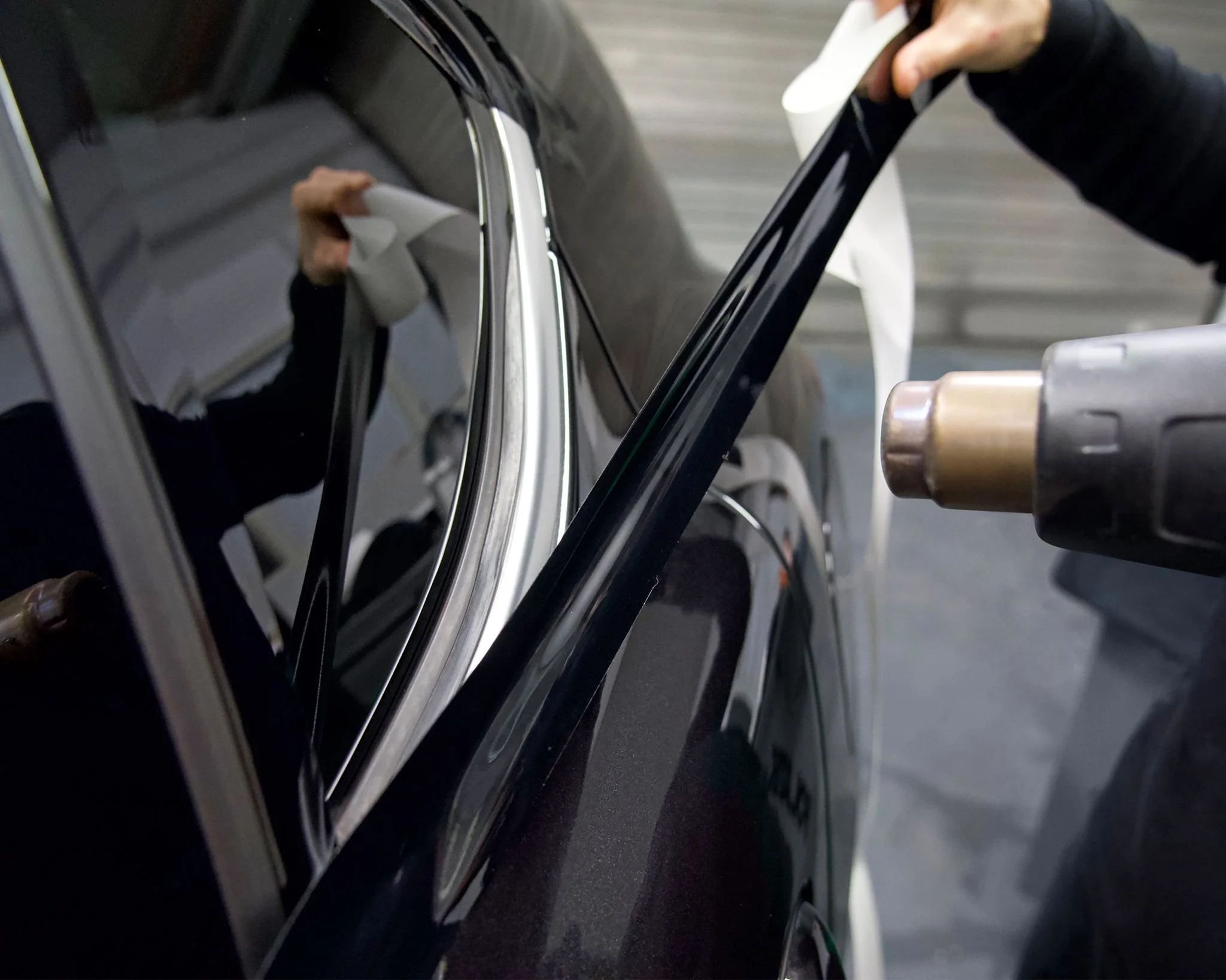 A person is using a heat gun to heat the black door trim of a black car, which is reflected in the car's side mirror.