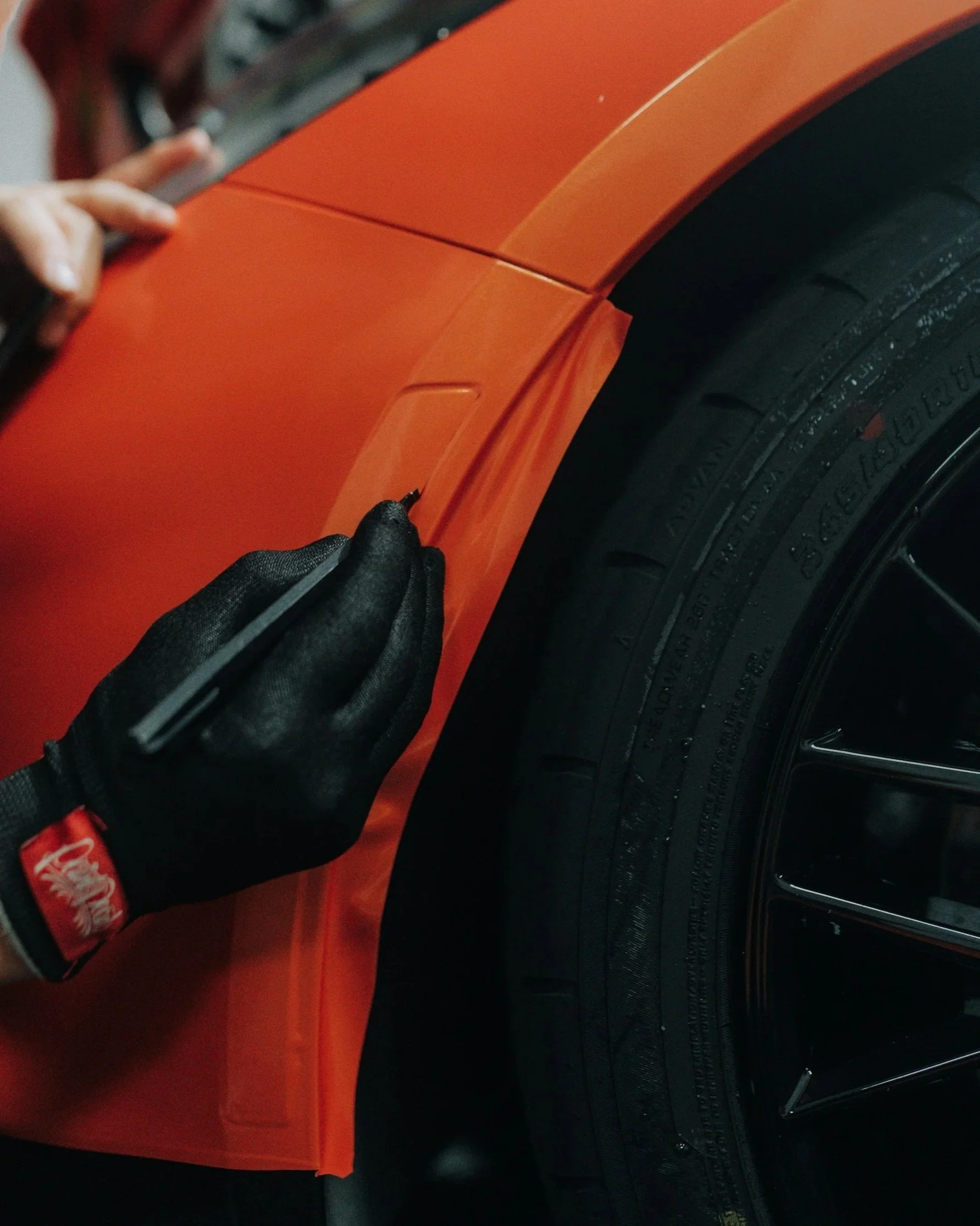 A person wearing a black glove is marking a small rectangular area on the orange body of a car with a black marker, next to a black tire.