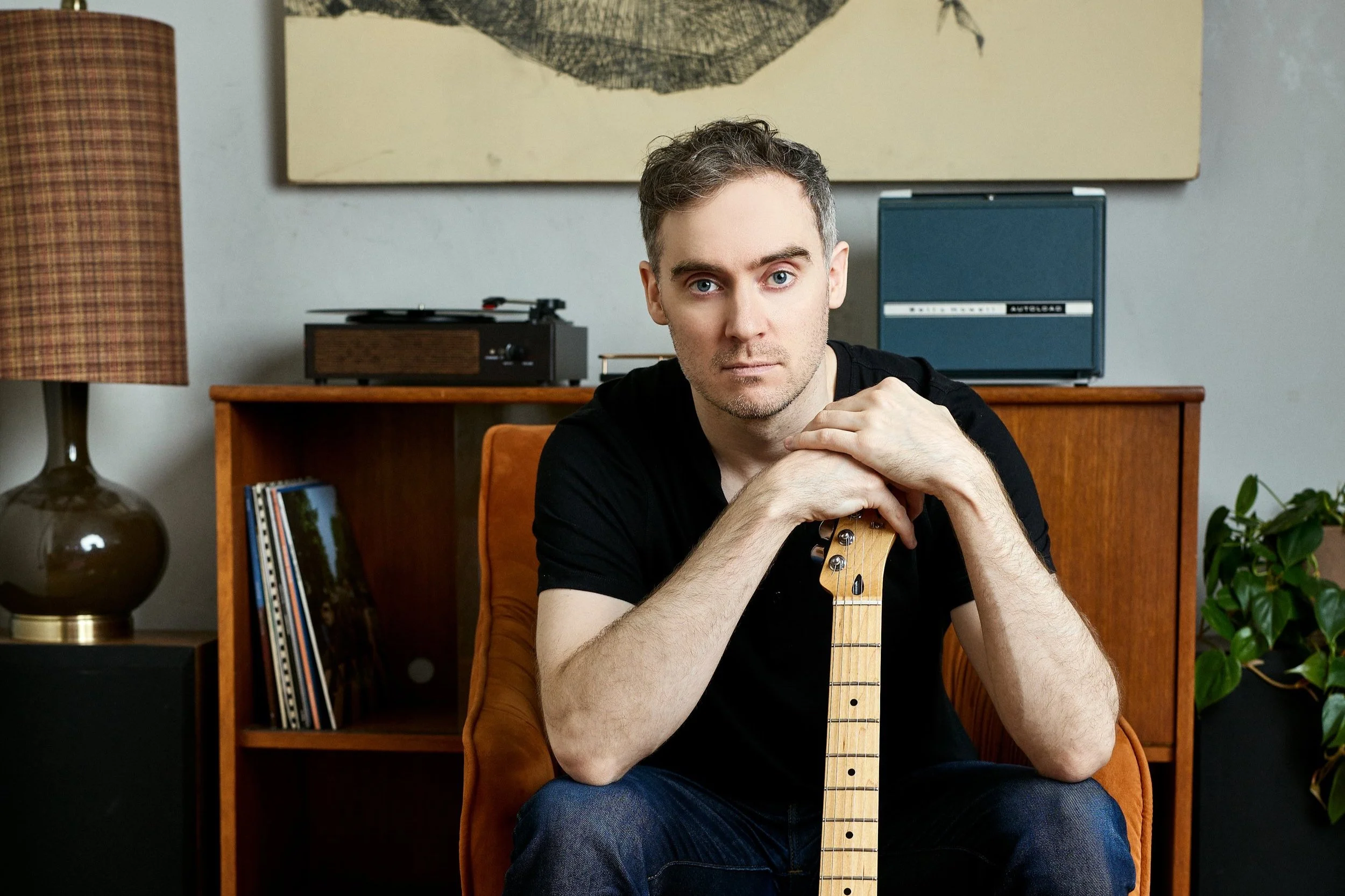 A man sitting on an orange velvet chair, holding a guitar neck vertically, with a serious expression, in a room with a wooden cabinet, vinyl records, a vintage record player, a lamp, and a small speaker, with modern art hanging on the wall behind him.