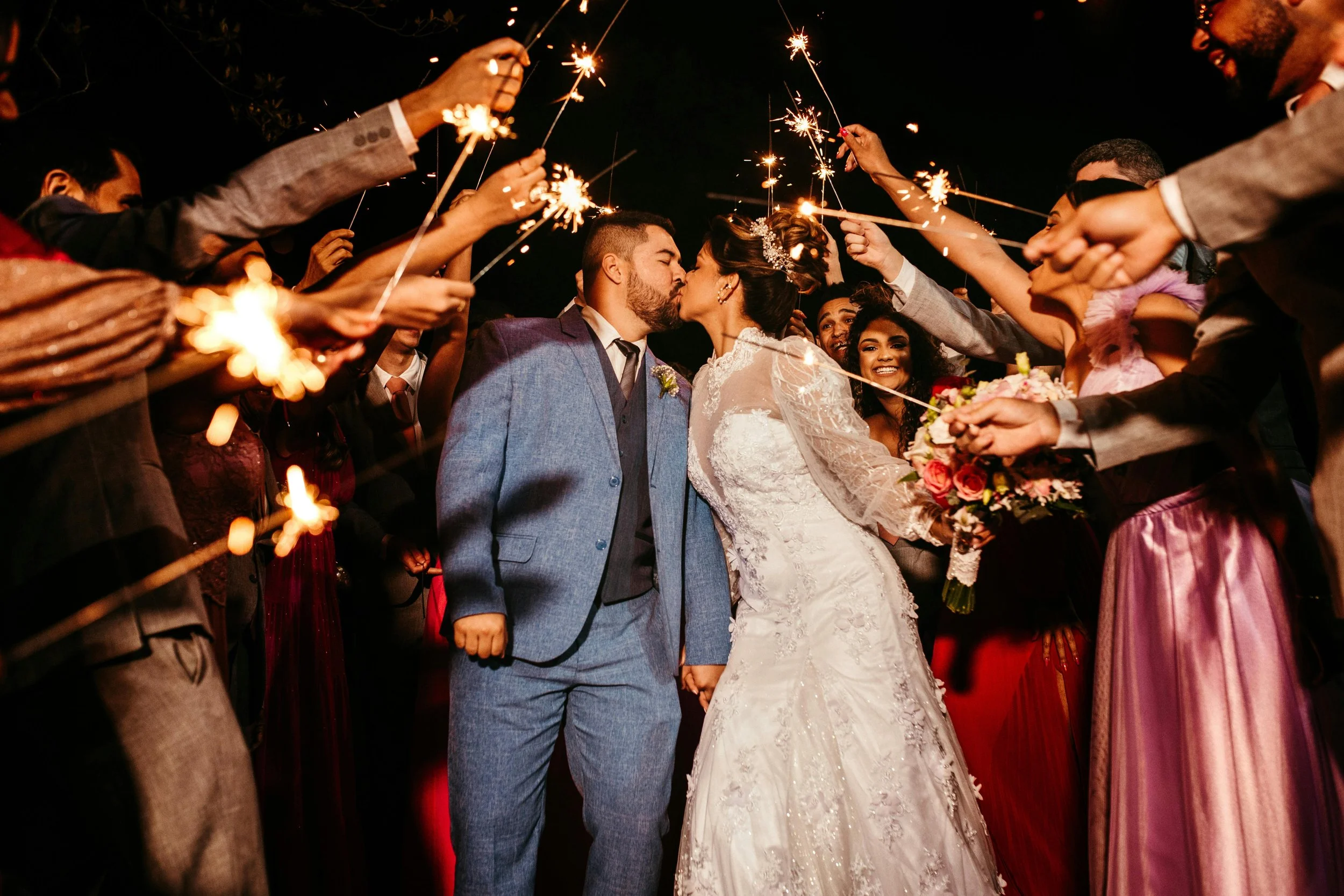 A wedding couple sharing a kiss surrounded by friends and family holding sparklers at night.