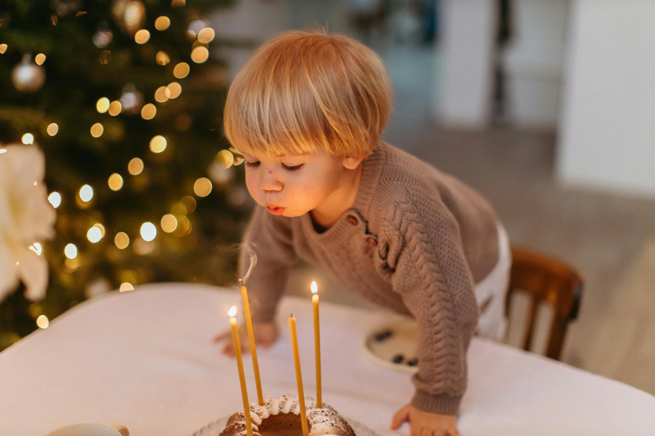 A young child with blond hair blowing out candles on a birthday cake during a celebration with a Christmas tree in the background.