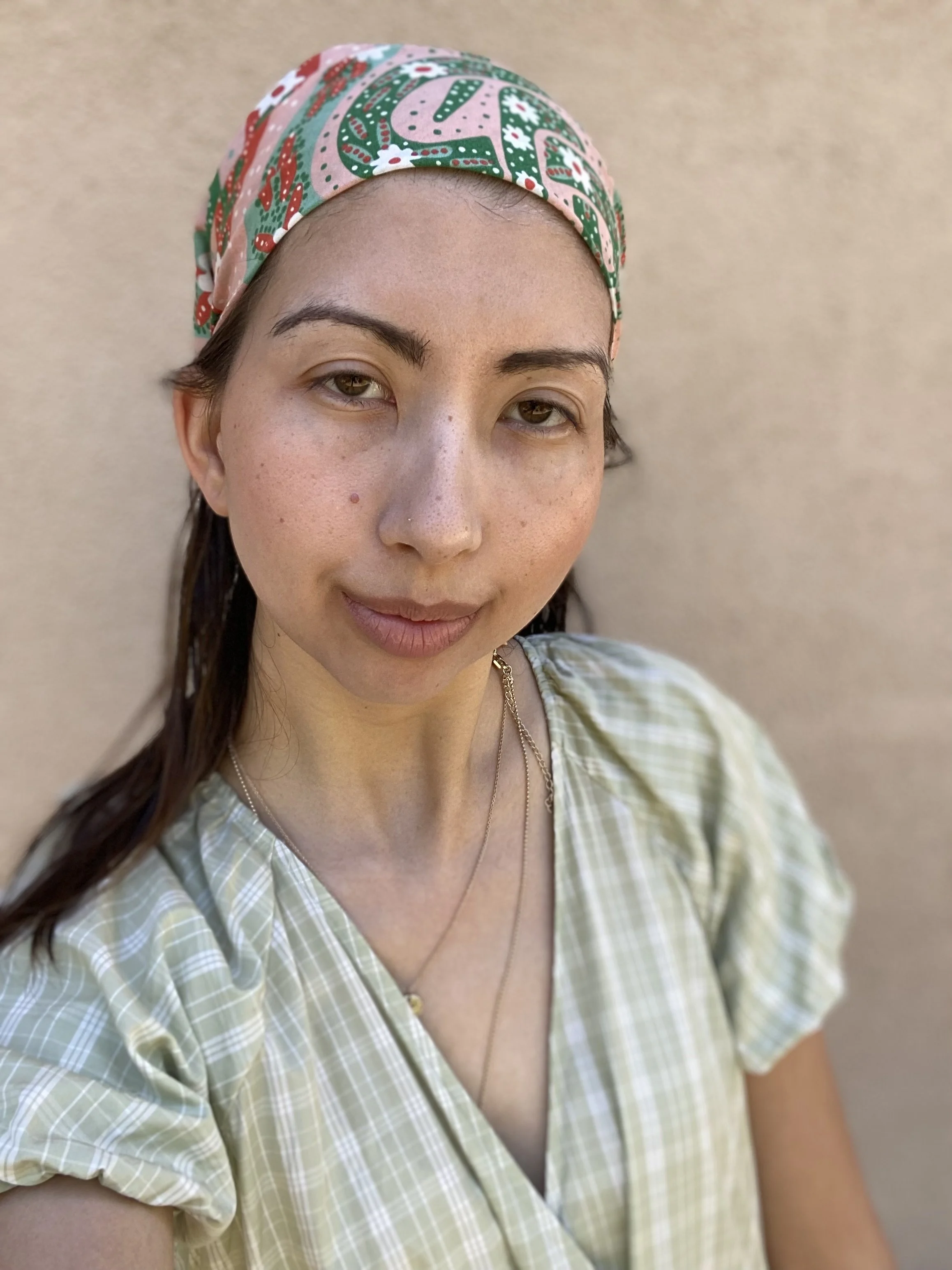 A young woman with light skin, brown eyes, and dark brown hair wearing a colorful patterned headscarf and a light green checkered blouse, standing against a beige wall.