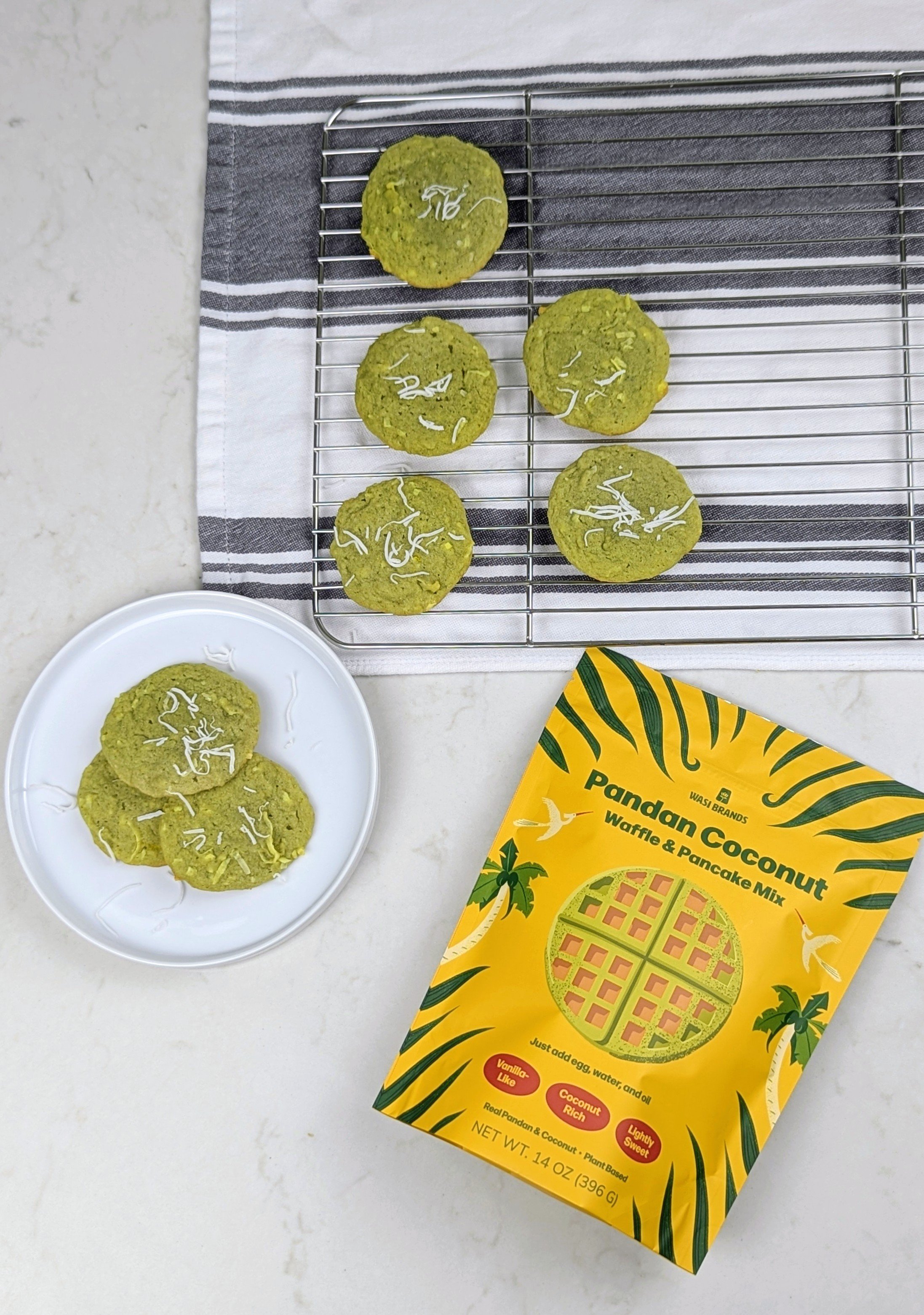 Green pandan coconut cookies with shredded coconut topping on a cooling rack and a bowl of cookies on a white surface, with a yellow pandan coconut waffle and pancake mix package.