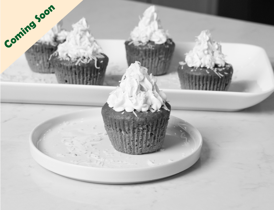 A close-up of a chocolate cupcake with white frosting on top, placed on a white plate. Four more cupcakes with white frosting are visible in the background on a rectangular tray, with a 'Coming Soon' banner in beige and green in the top left corner.