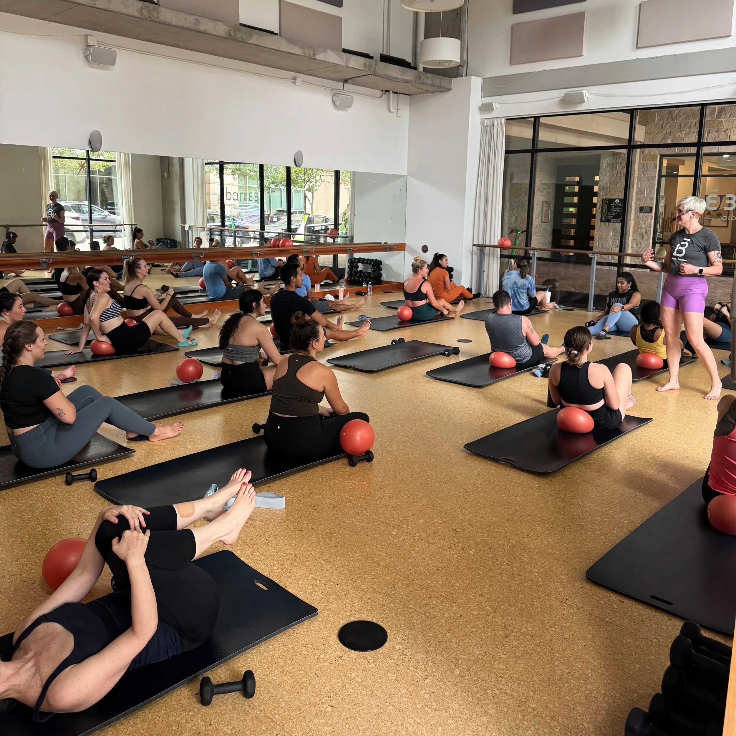Women participating in a fitness class at a gym, seated on mats with exercise balls, instructor leading the class. Large windows with a view outside, mirrored wall, ceiling fans, and modern decor inside the gym.