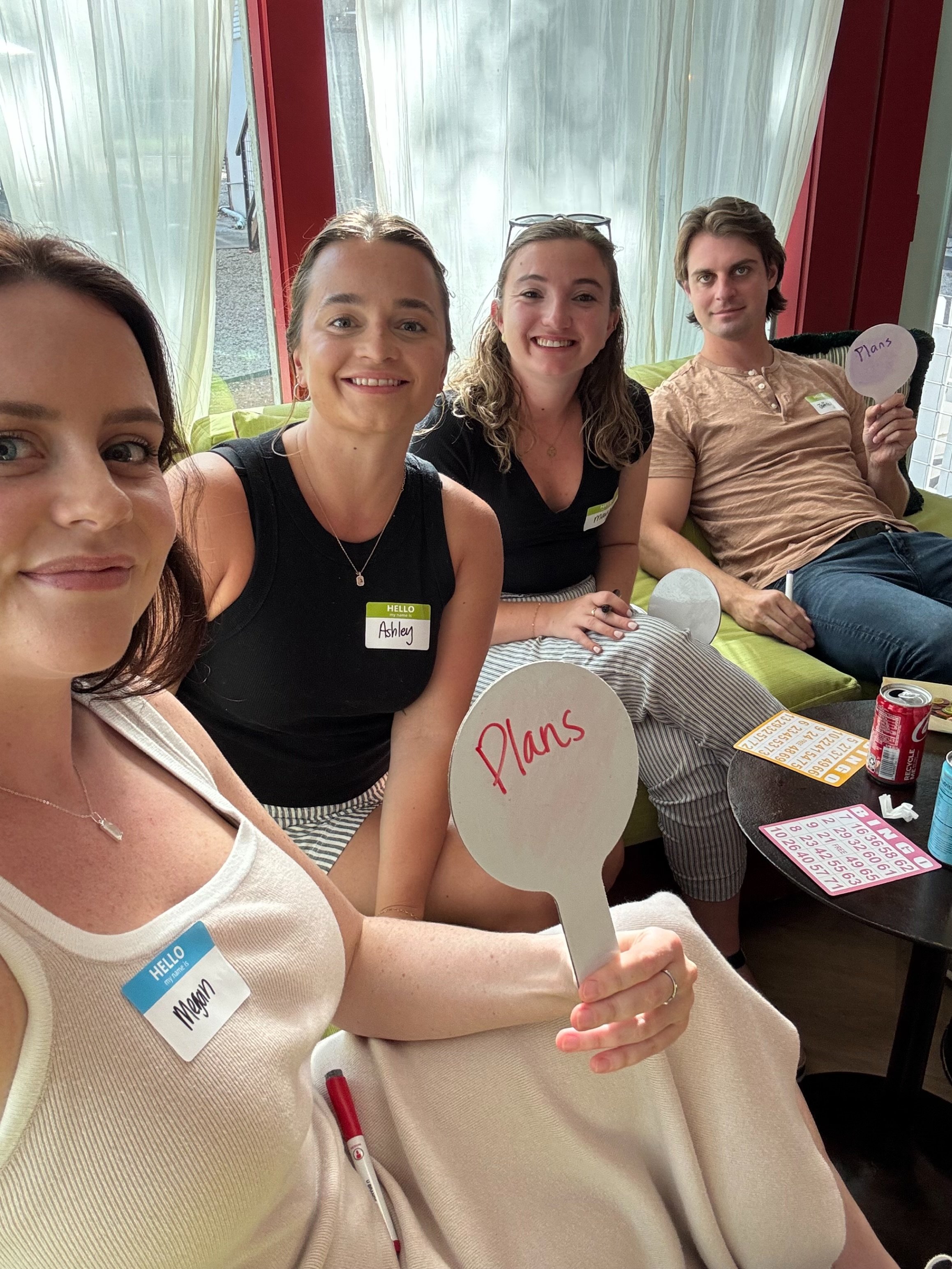 Group of four smiling people sitting on a couch during a social gathering or game night, with a table nearby holding bingo cards, a soda can, and bingo plan signs. The woman in the foreground, wearing a beige dress and a name tag that says "Megan," i