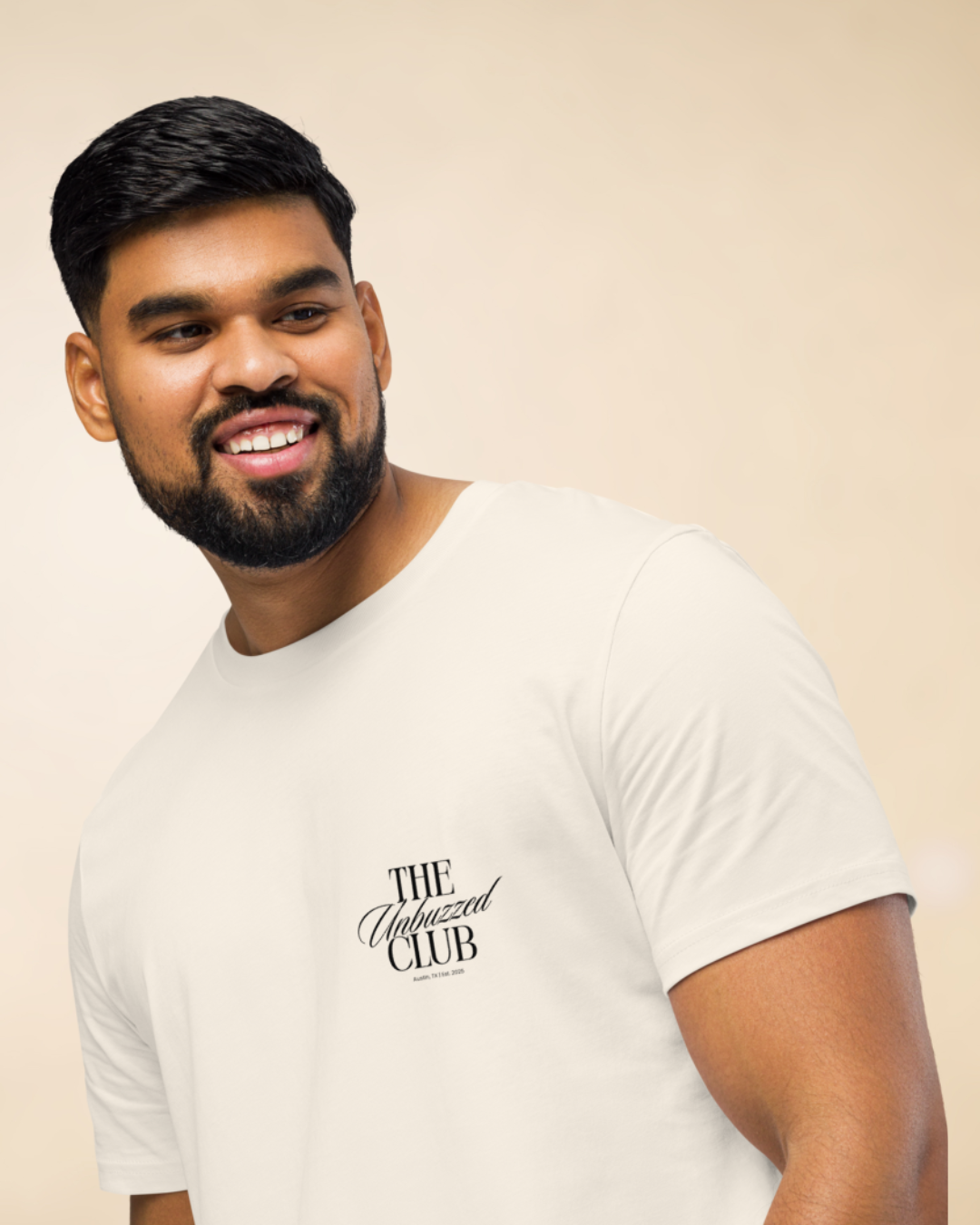 A man with a beard and short black hair wearing a white T-shirt that says 'THE Unbuzzed CLUB' smiling and looking to his left.