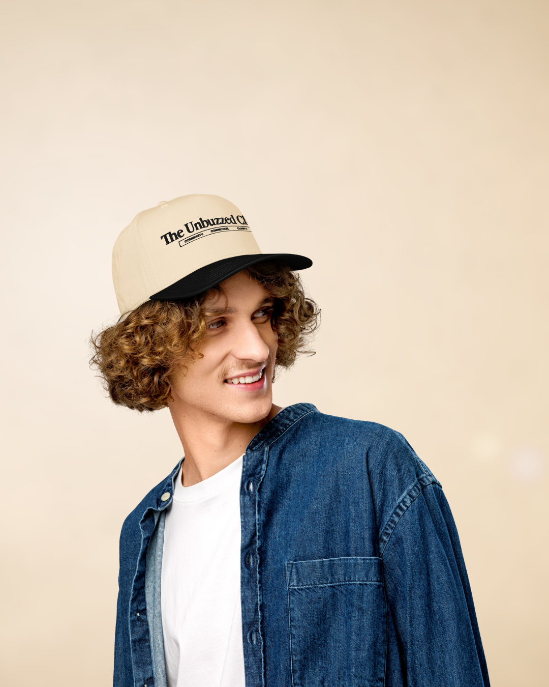 A young man with curly hair wearing a baseball cap, a white T-shirt, and a denim jacket, looking to his left with a smile.