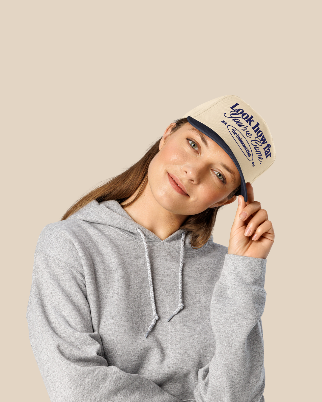 Young woman wearing a gray hoodie and a beige cap with blue text, tilting her head and holding the cap's brim, on a plain beige background.
