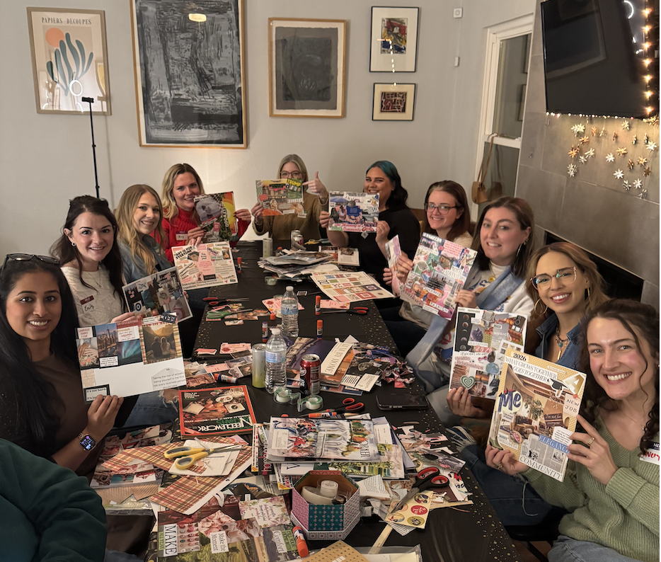 Group of women gathered around a large table, engaged in crafting with magazines, scissors, glue, and other supplies, smiling and showing off their completed projects in a cozy, decorated room.