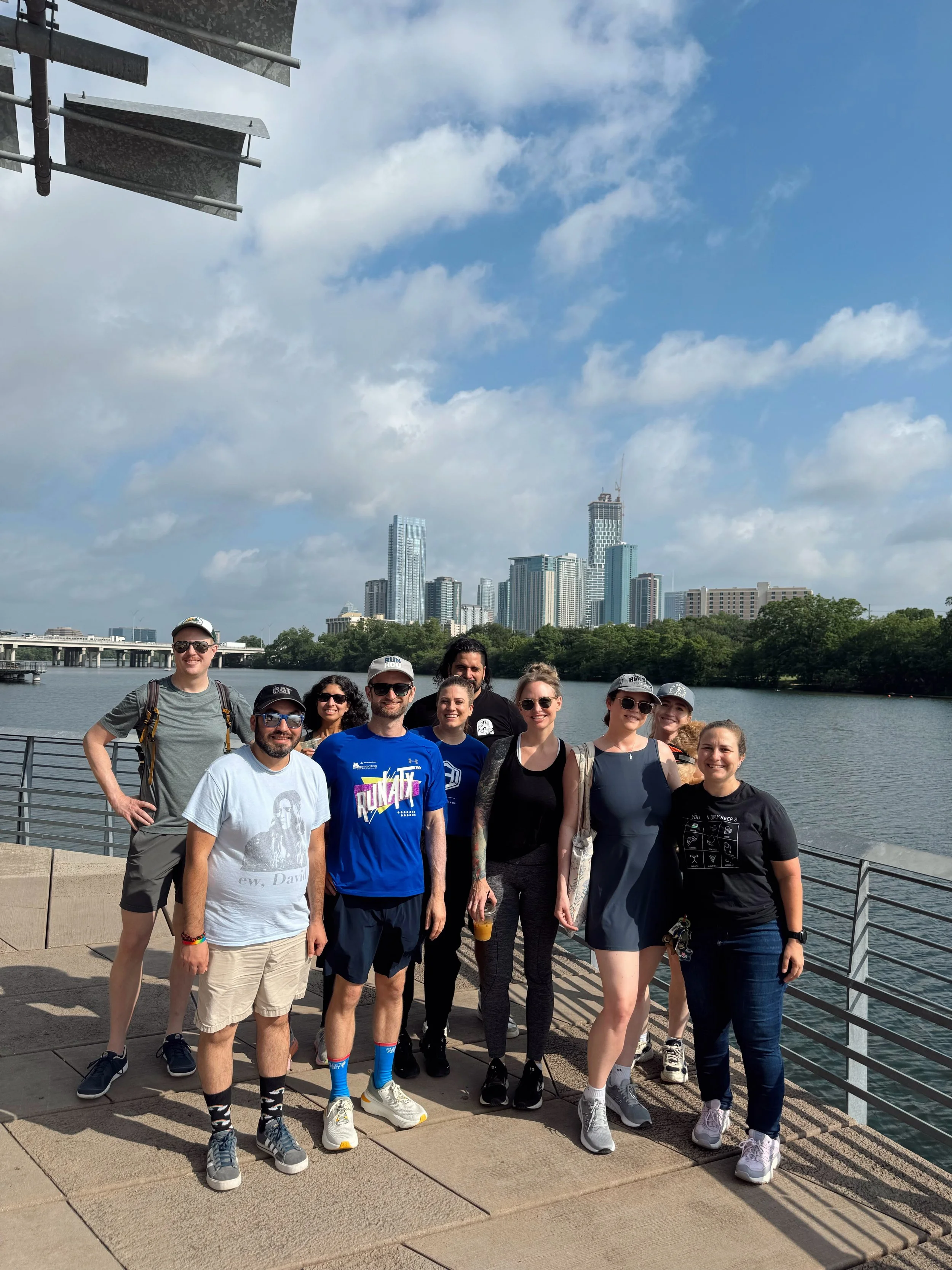 A group of ten people standing on a boat dock, smiling with a river and a city skyline in the background under a partly cloudy sky.