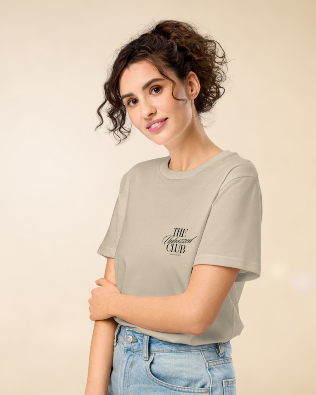 A young woman with curly brown hair and light skin is wearing a beige t-shirt with black text that reads 'THE Unfuzzed CLUB' and light blue jeans. She is standing against a plain light background, smiling softly, with her arms crossed.