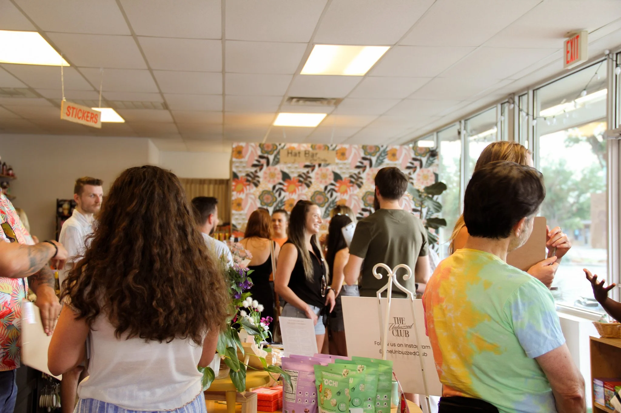 People inside a store or cafe, some are browsing products, and others are talking, with a woman at the counter and a floral patterned wall in the background.