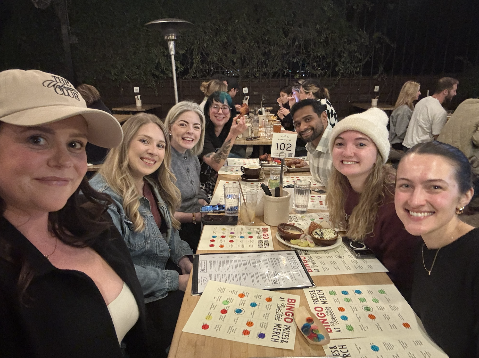 Interior of a restaurant or cafe with a long table filled with people engaged in conversations, brick walls, large windows, hanging lights, and a bar area on the right side.