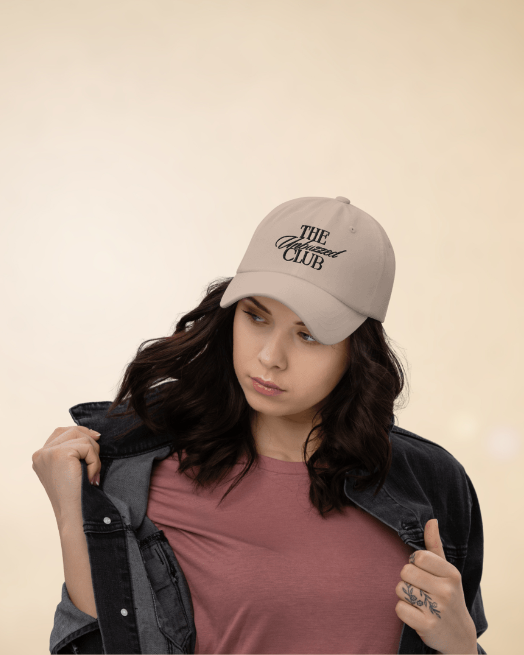 Woman wearing a beige baseball cap with 'The Unhoused Club' written on it, a pink t-shirt, and a black denim jacket, looking down and to the side against a plain background.