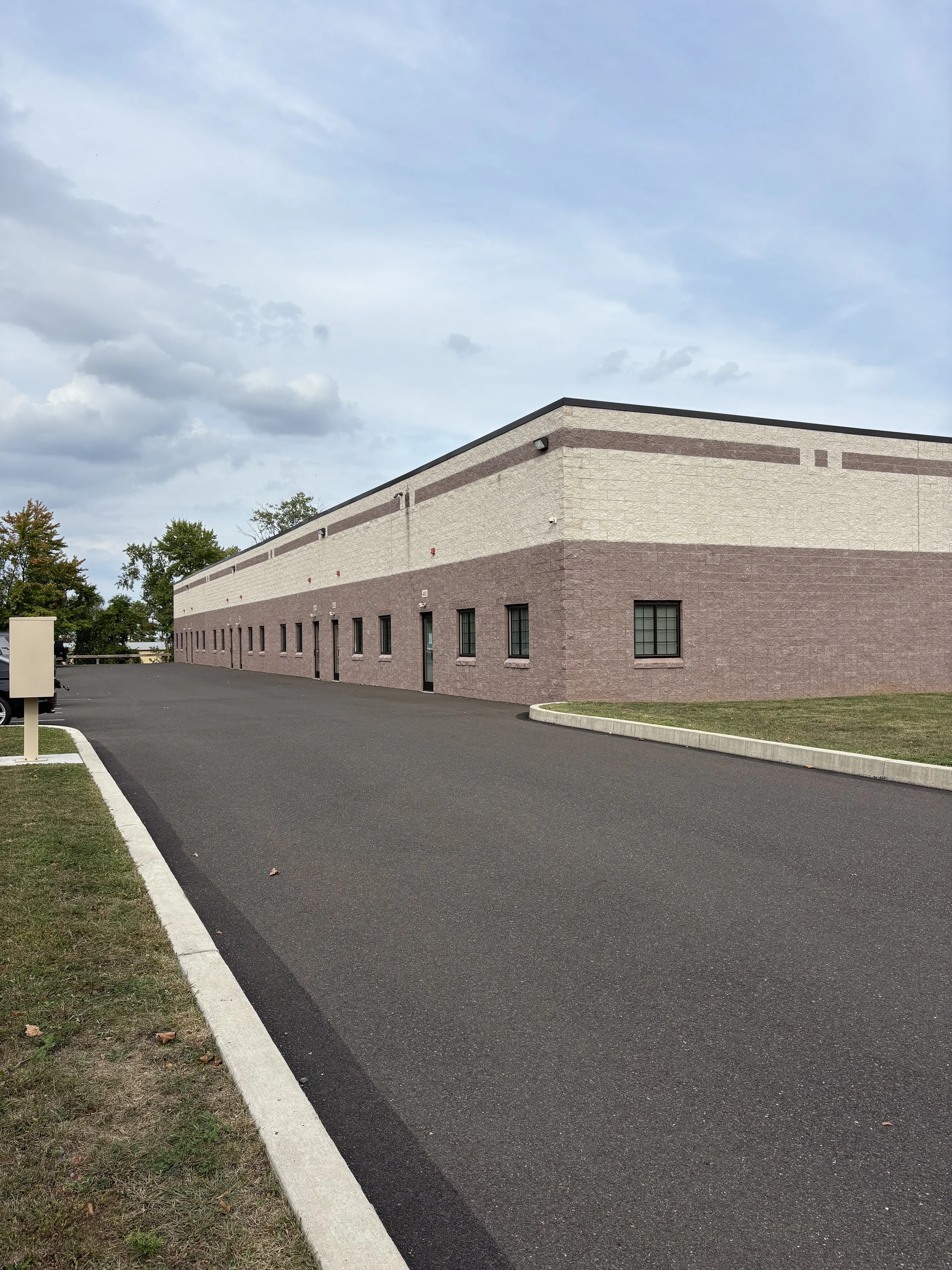 Exterior view of a large industrial brick warehouse with multiple windows, parked cars, a paved parking lot in front, and a partly cloudy sky.