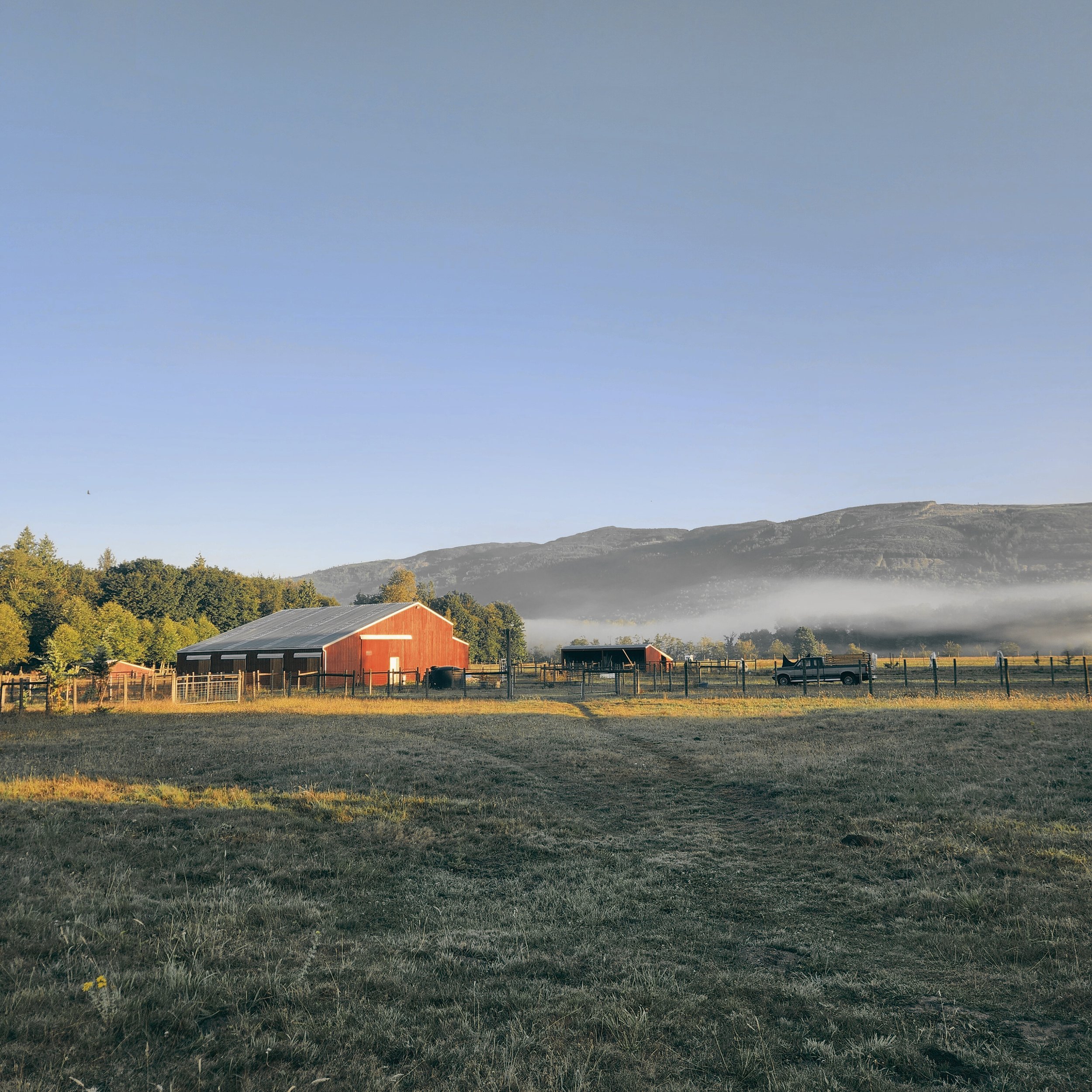 A rural farm scene with a red barn, a smaller shed, a truck, a wooden fence, trees, and distant hills with low-lying fog and a clear blue sky.