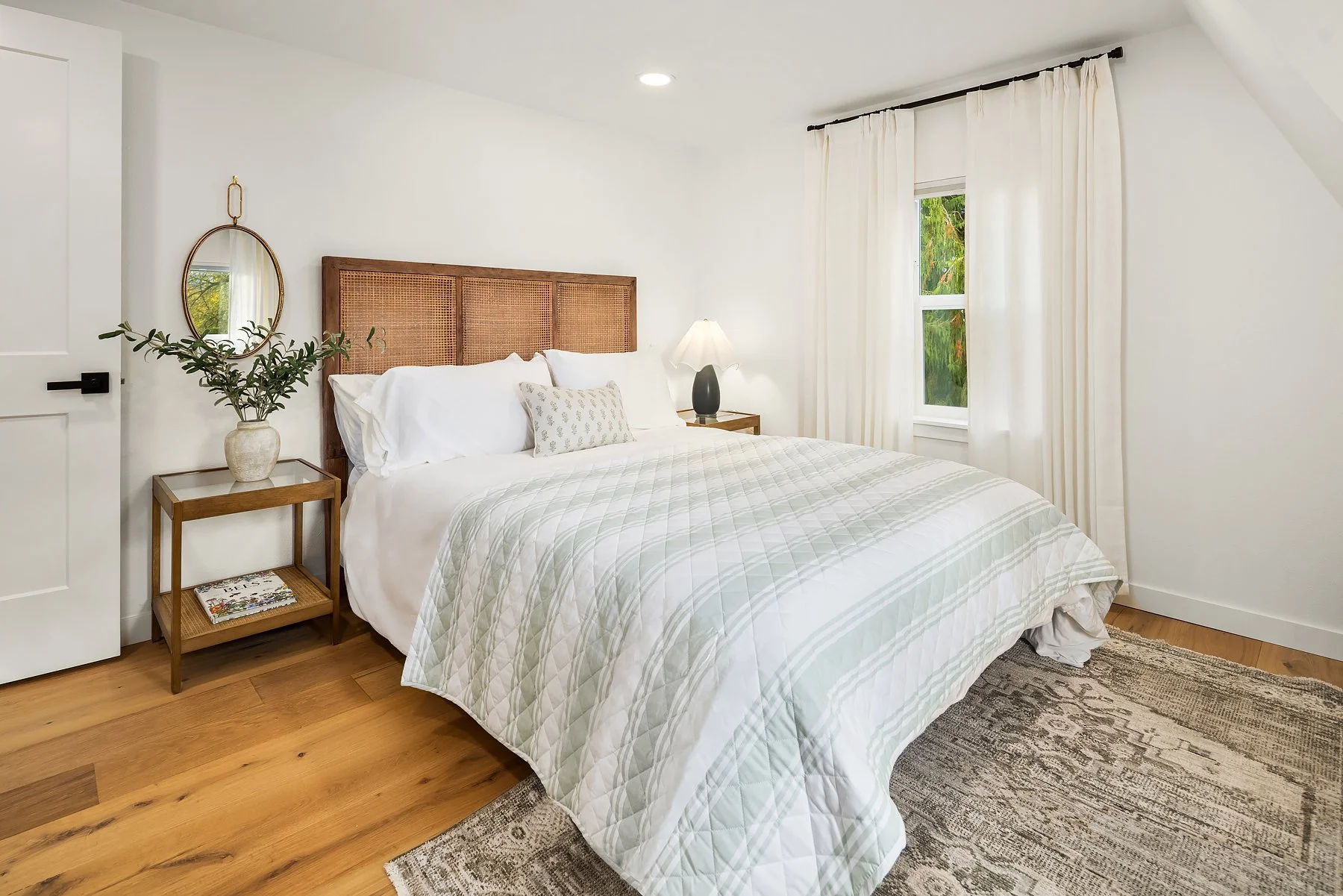 A cozy bedroom with a large bed that has white bedding and pillows, wooden side tables with a lamp and a potted plant, white curtains on a window, and hardwood floors.