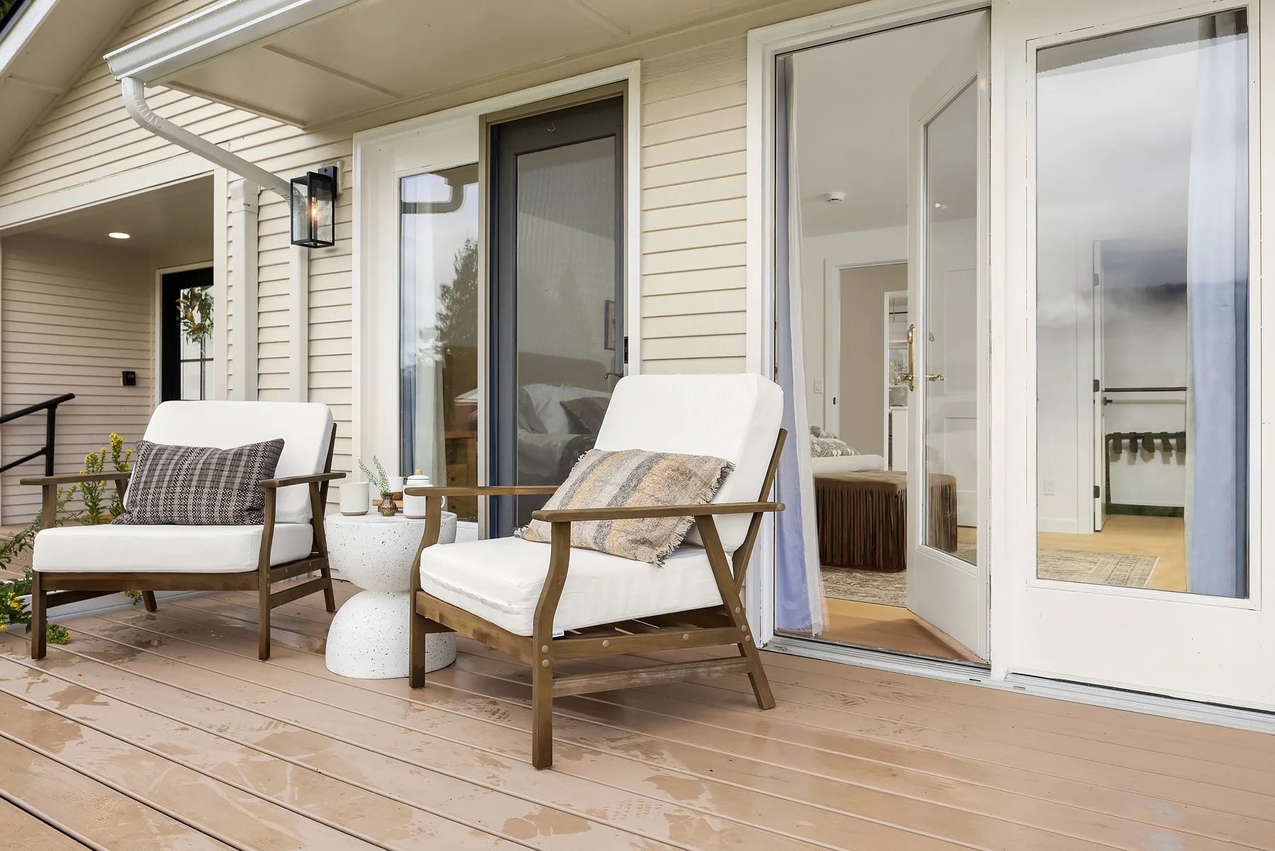 A wooden deck with two white cushioned chairs and a small white speckled side table outside a house with sliding glass doors. The interior of the house is visible through the open door.