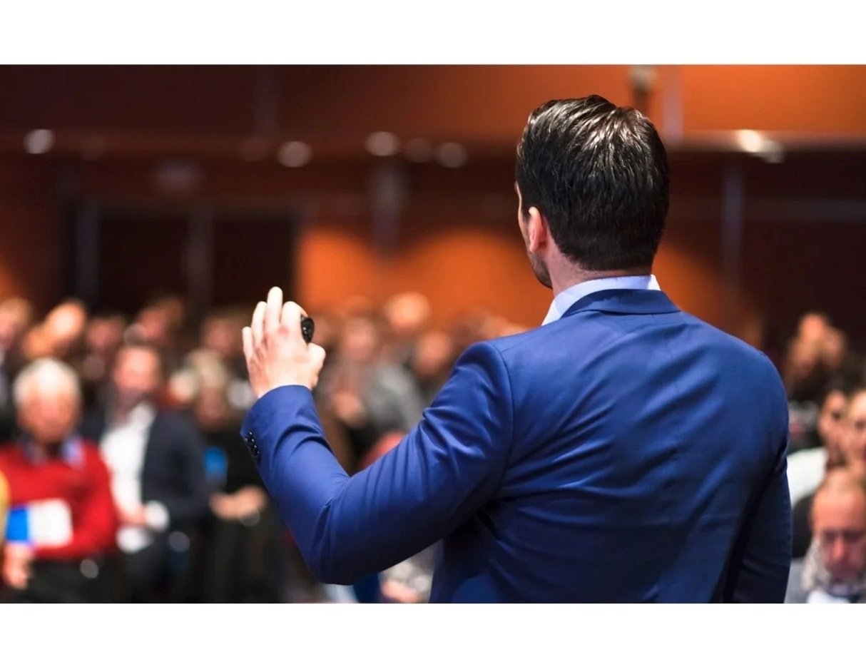 A man in a blue suit holding a microphone in front of an audience at a conference or seminar.