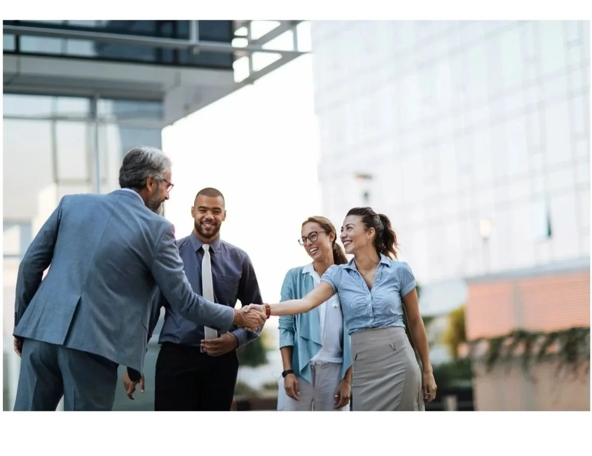 A group of four business professionals, two men and two women, shaking hands outside a modern office building, smiling and greeting each other.