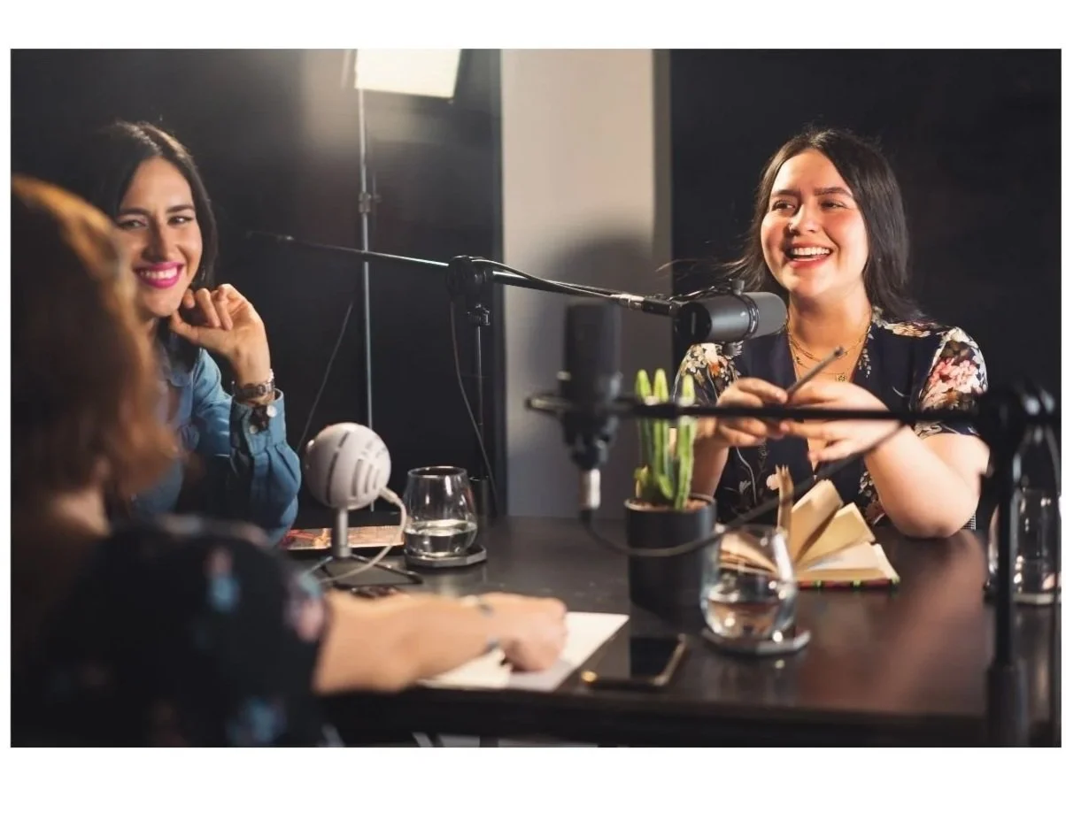 Three women participating in a podcast recording session, with one woman speaking into a microphone and two women listening and smiling at the table with notebooks, glasses of water, and a potted plant.