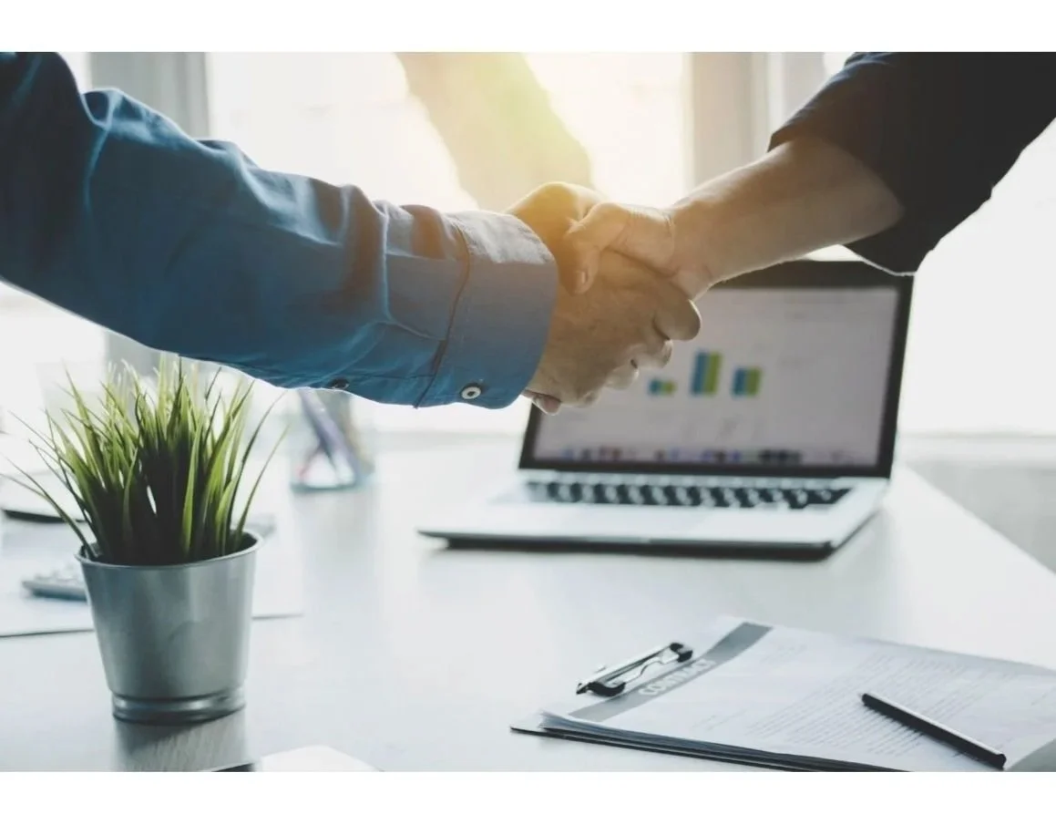 Two people shaking hands in an office, with a laptop showing graphs, a clipboard, and a potted plant on the desk.