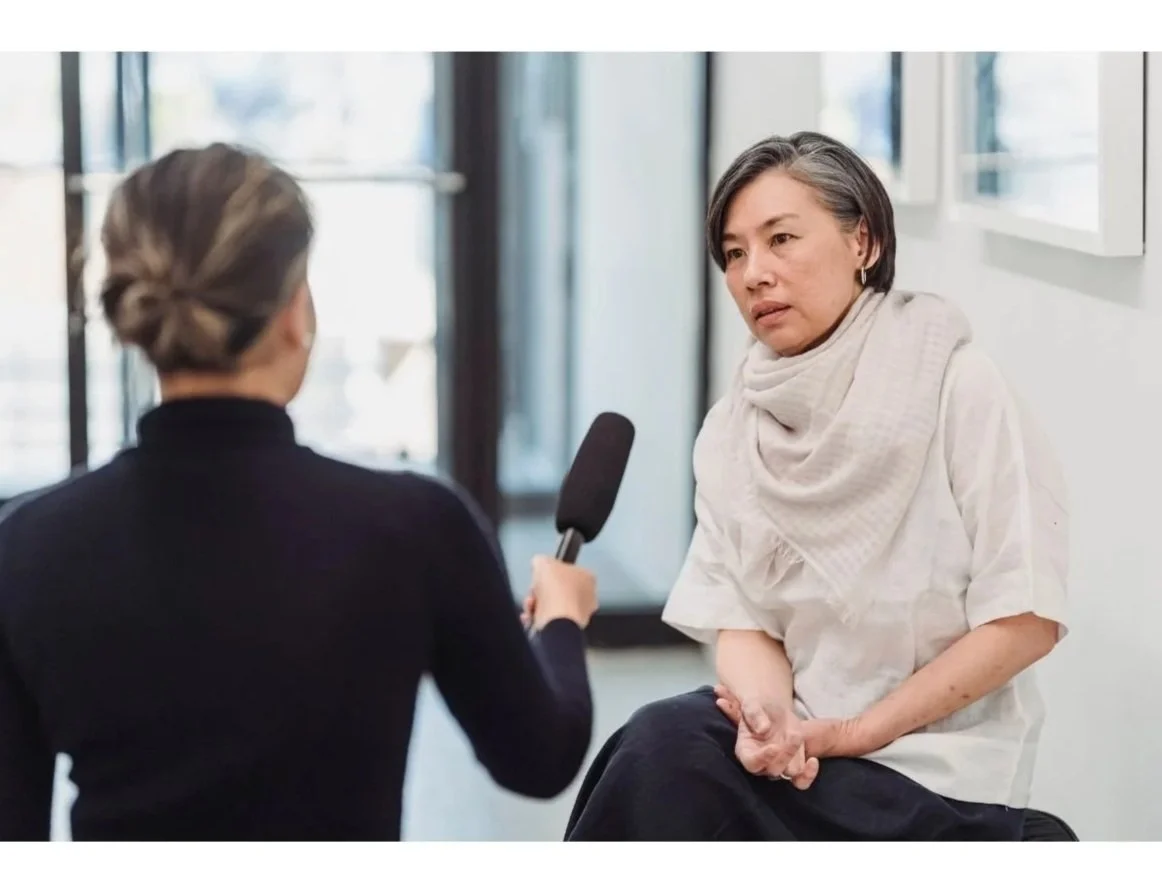 An interview scene showing a woman with short grey hair and a white shirt talking to a reporter holding a microphone in an indoor setting.