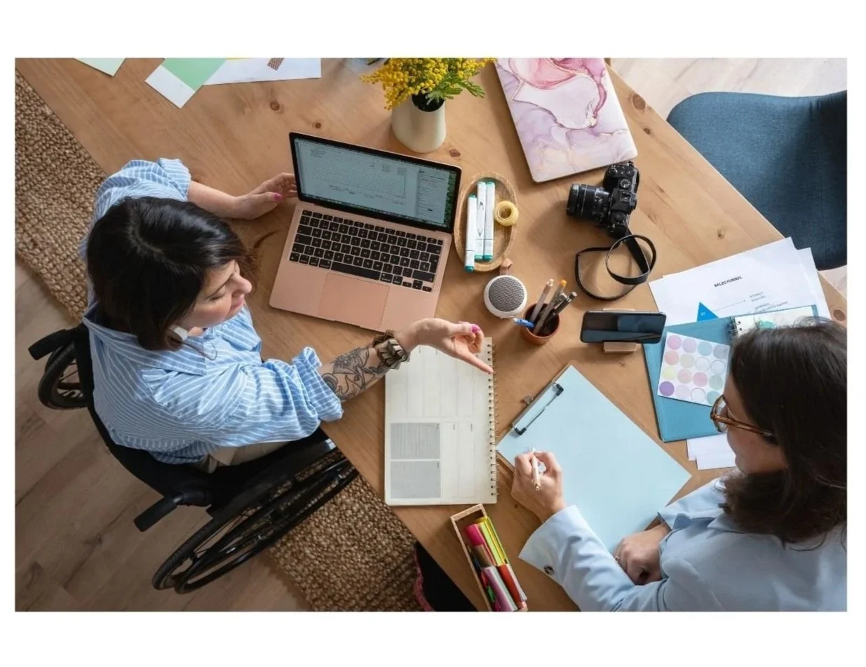Two women working at a wooden table with laptops, notebooks, camera, and art supplies, viewed from above.