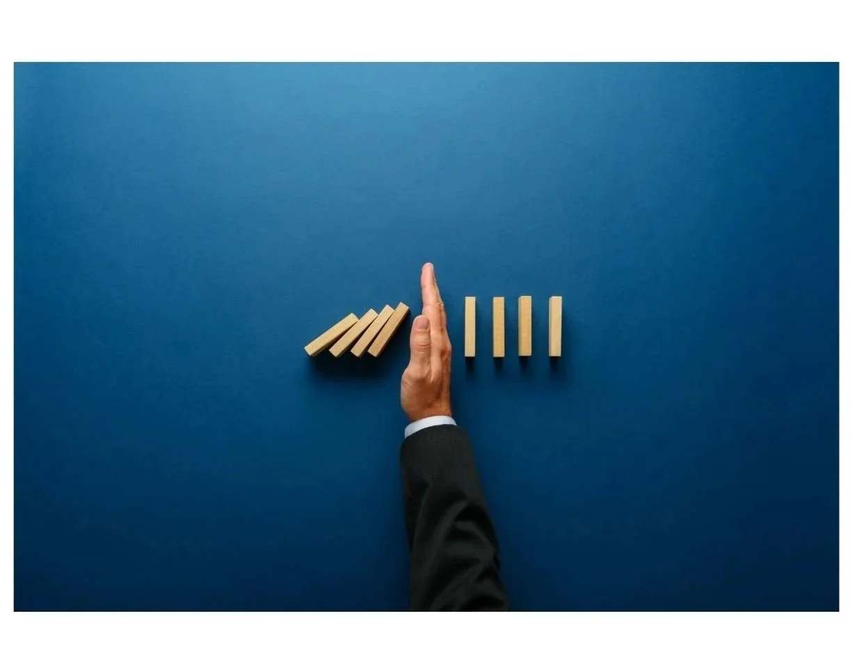 A person in a business suit is stopping a line of wooden dominoes from falling with their hand on a blue background.