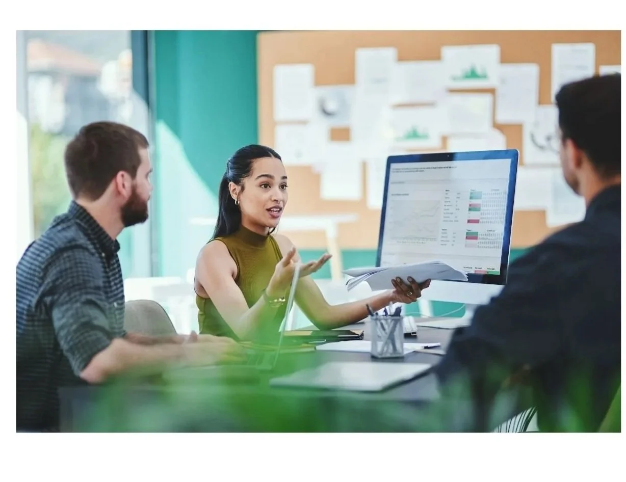 Three people having a business meeting in an office, with one woman explaining data on a computer monitor to two men.