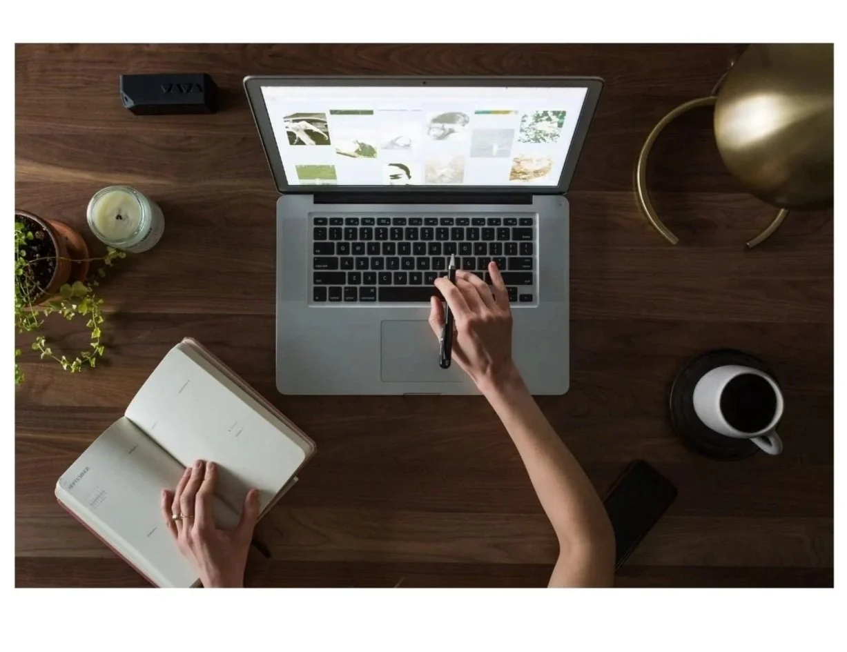Top-down view of a wooden desk with a person using a laptop and holding a pen, an open notebook, a smartphone, a coffee mug, a candle, a potted plant, a gold desk lamp, and a black speaker.