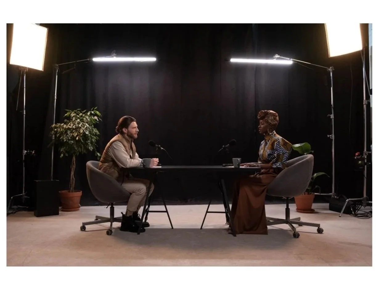 Two people participating in an interview or podcast recording in a studio with black curtains, studio lights, and potted plants.