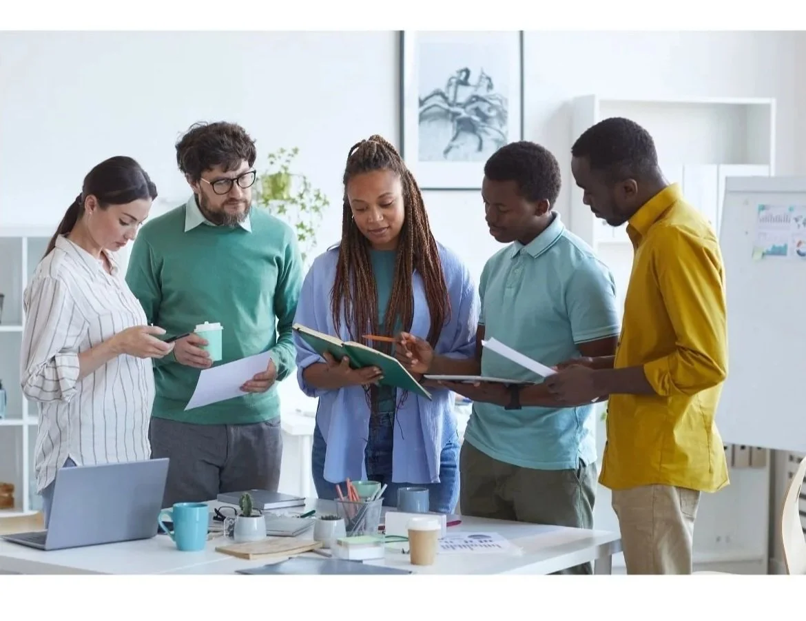 A diverse group of five people gathered around a table in an office, looking at documents and discussing.
